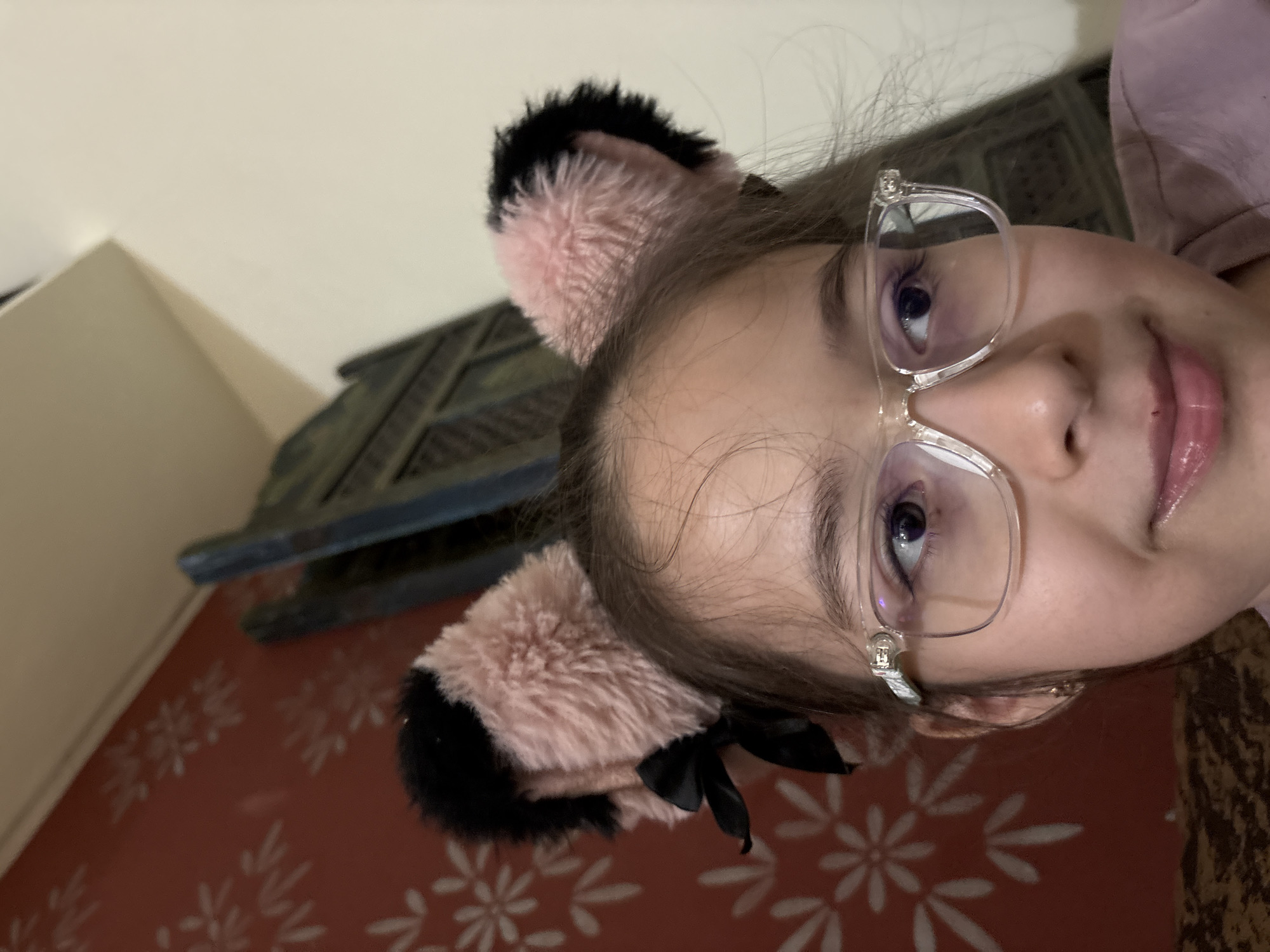 A close-up, head-and-shoulders shot shows a young person wearing clear-rimmed glasses and furry, pink and black cat ears. The person is looking to their left with a slight smile. A portion of their face and the faux ears are illuminated by the camera's flash. The background is indistinct, with hints of a patterned red wallpaper and what appears to be a dark, ornate piece of furniture. The image was taken indoors, possibly in a residential setting in Ashvem, India.