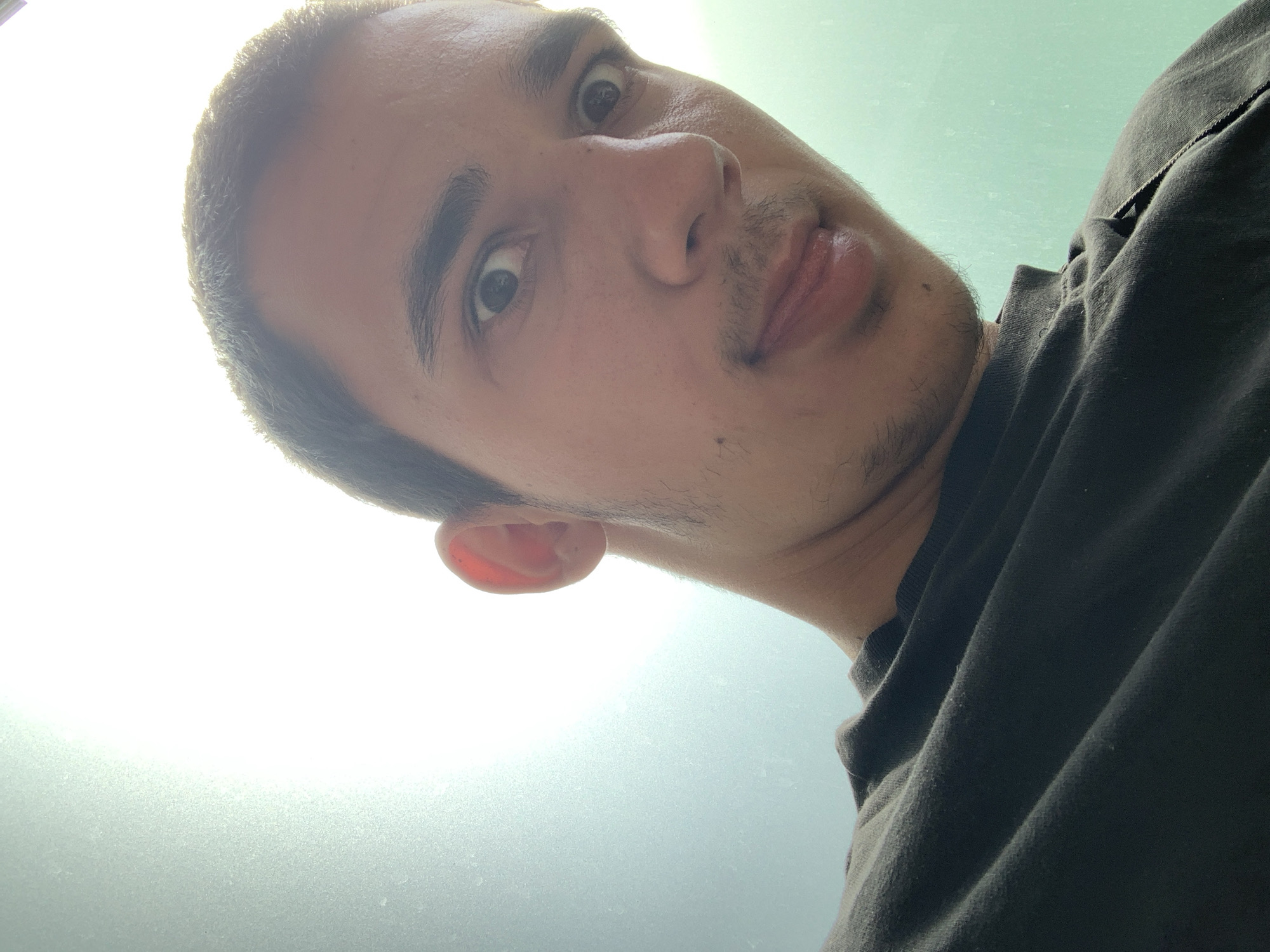 A selfie taken from a low angle looking up at a man's face. He has short, dark hair, light skin, and dark eyes. He has a mustache and some stubble on his chin. He is wearing a dark t-shirt. The background is a light, unclear color, possibly from a window or light source. There is no indication that the photo was taken in Dundrum, Ireland or any other specific location.