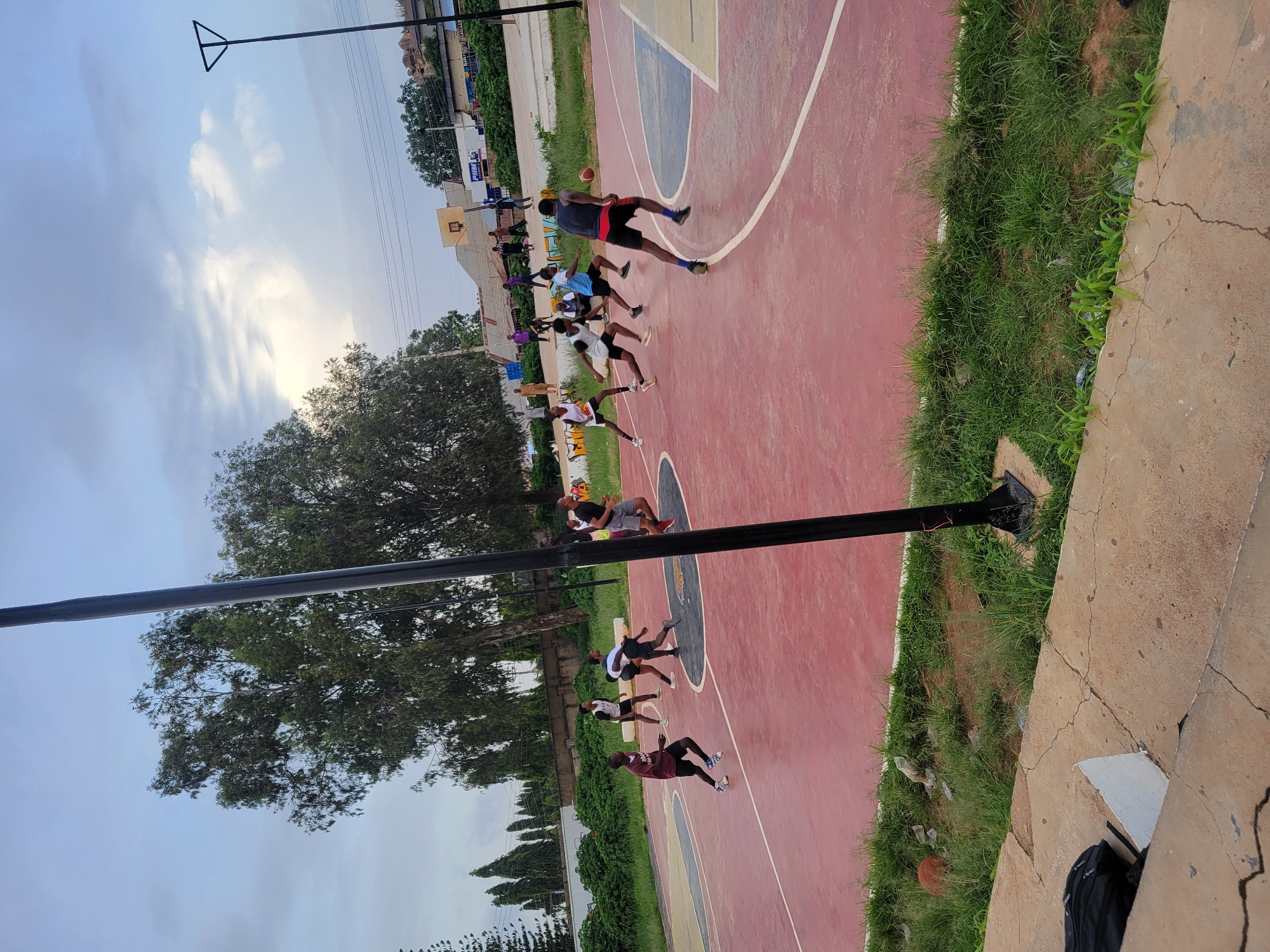 In the image, a group of young men are engaged in a basketball practice session on an outdoor court. The court has a red playing surface with white lines marking the boundaries and key areas. The game appears to be in progress, with one person dribbling a basketball, while others are in various stances, suggesting they are either playing defense or preparing to receive a pass. The time of day seems to be late afternoon or early evening, as indicated by the soft, diffused light and the sky, which shows a mix of clouds and sunlight. The weather appears to be pleasant, with clear skies and greenery surrounding the court.
The subjects are dressed in athletic wear, mostly shorts and t-shirts. One individual stands out with a dark grey tank top and red shorts, actively playing with the ball. Another person in a white t-shirt and black shorts is positioned in front of him, likely playing defense. Further down the court, several other individuals are visible, some standing and others in motion.
The setting includes a basketball hoop and backboard at one end of the court, though not fully visible. Lush green grass borders the court, and trees are seen in the background, suggesting an outdoor recreational area. The presence of a tall pole with a triangular fixture overhead might indicate lighting for the court or a utility pole.
Notable details include a basketball on the ground near the edge of the court, and a black backpack placed on some concrete steps. The overall mood seems energetic and focused, typical of a sports practice. There is no visible text in the image that provides specific information about the location or event.