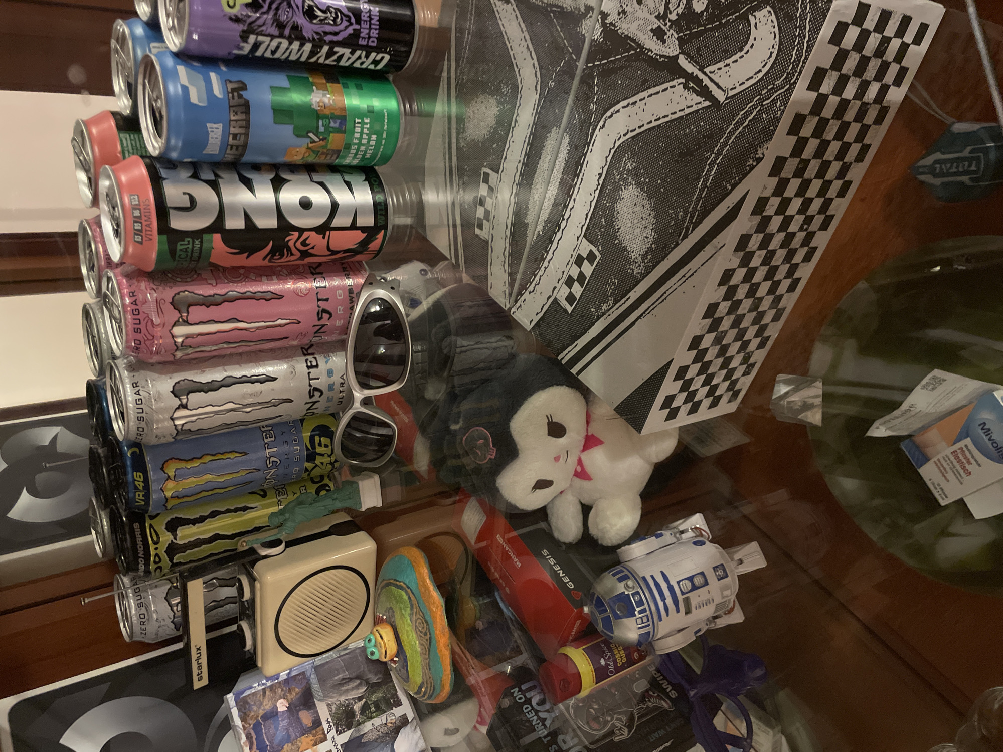 The image shows a collection of objects on a glass table, likely in a room with wood furniture. The objects include stacked energy drink cans (brands like Monster, Kong, and Crazy Wolf), a shoe box with a checkerboard pattern and a shoe design, sunglasses, a plush toy, a small radio, a miniature R2-D2 figure, a red tube labeled "MANGAN", and other small items. The text "CAN'T WAIT" and "YOUR HAR" is visible on an object. The objects are clustered together, creating a somewhat chaotic arrangement. There is no information regarding the city of Sofia, Bulgaria.