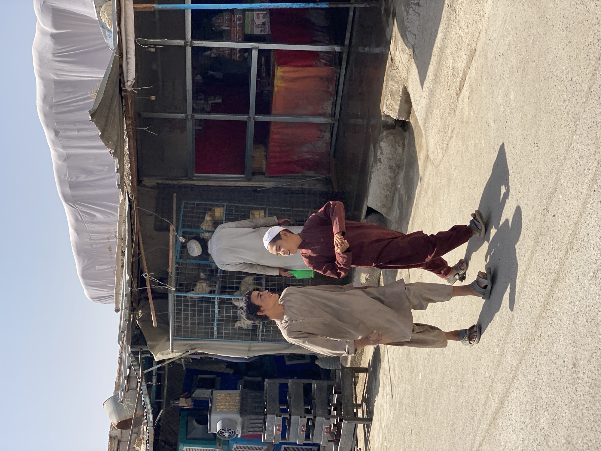 In Kandahar, Afghanistan, two young boys, one in a tan tunic and the other in a maroon tunic, walk down a paved street under a bright sky. They are engaged in conversation, their faces turned towards each other. In the background, a man in a white tunic and a white skullcap walks away from the camera. He is near a building with a corrugated metal roof covered by a large white tarp. The building appears to be a market stall or shop, with various goods visible through a metal-grated enclosure. Stacks of what appear to be gas stoves or similar equipment are to the left of the boys. The ground is uneven, with concrete curbs and patches of dirt. The overall scene suggests a typical street in a bustling Afghan city.