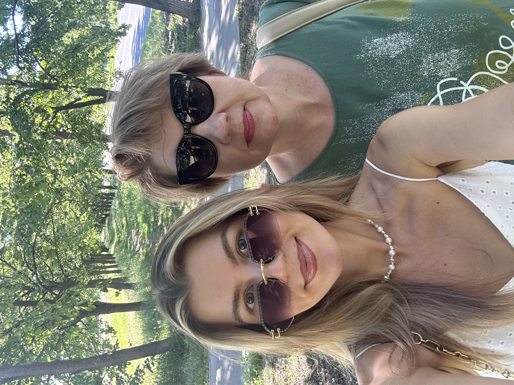 Two women are posing for a selfie. The woman in the foreground is younger, with blonde hair and wearing sunglasses and a pearl necklace. She is wearing a white top. Behind her is an older woman with short blonde hair, also wearing sunglasses and a green shirt. The background shows a green outdoor scene with trees and a path, suggesting they are in a park or similar green space. The location is Unknown, Unknown.