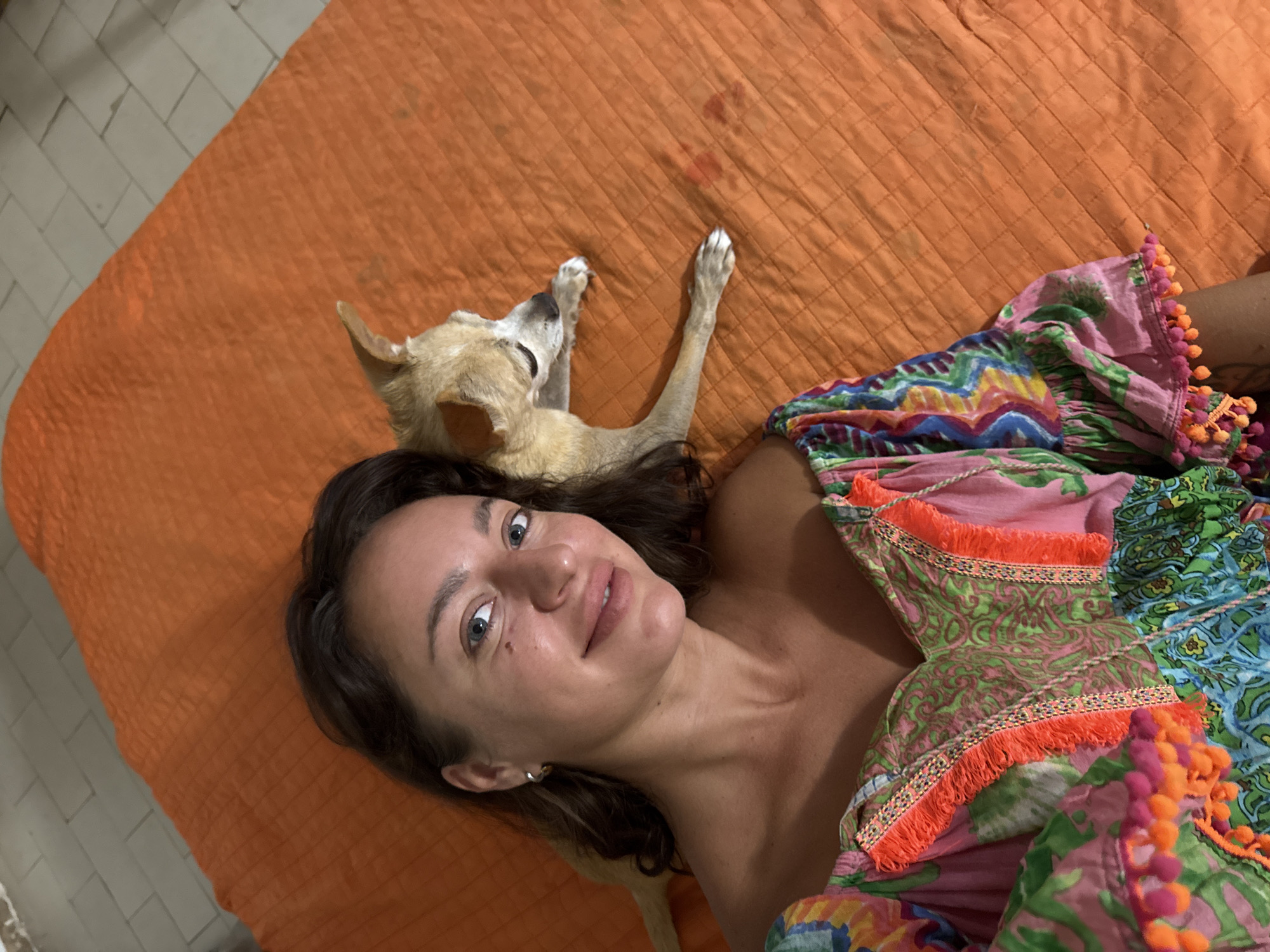 This media file shows a woman and a dog lying on an orange quilted surface, possibly a bed or a mat. The woman is lying on her back, with her head turned towards the camera, smiling softly. She has long, dark hair and is wearing a colorful, patterned dress with orange and pink pom-pom trim. A small, light-colored dog is lying next to her, with its head resting on her shoulder. The dog's eyes are closed, and it appears to be sleeping.
The scene is indoors, with a tiled floor visible in the background. The lighting suggests it is daytime, possibly late afternoon or evening, given the soft light. The woman's expression is relaxed and content, and her connection with the dog appears affectionate. The overall mood of the image is peaceful and intimate. The location is identified as San Benedetto del Tronto, Italy, but there are no specific visual cues in the image to confirm this. No visible text is present in the image.