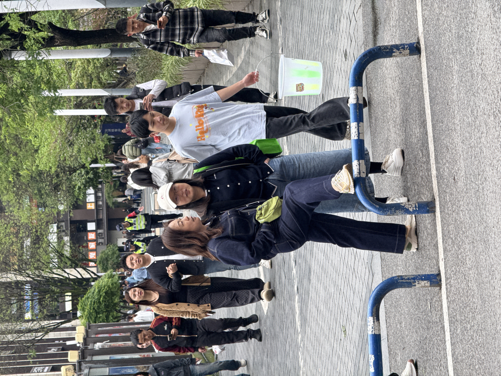 A daytime shot shows a crowd of people on a city street in Yuzhong District, China. The focus is on a group of people standing close to a blue metal barrier at the edge of a sidewalk. One individual is holding a translucent green bucket with a handle. Others are wearing casual clothing, including jeans, t-shirts, and jackets. The background shows buildings and more people, including a person wearing a bright yellow traffic vest.