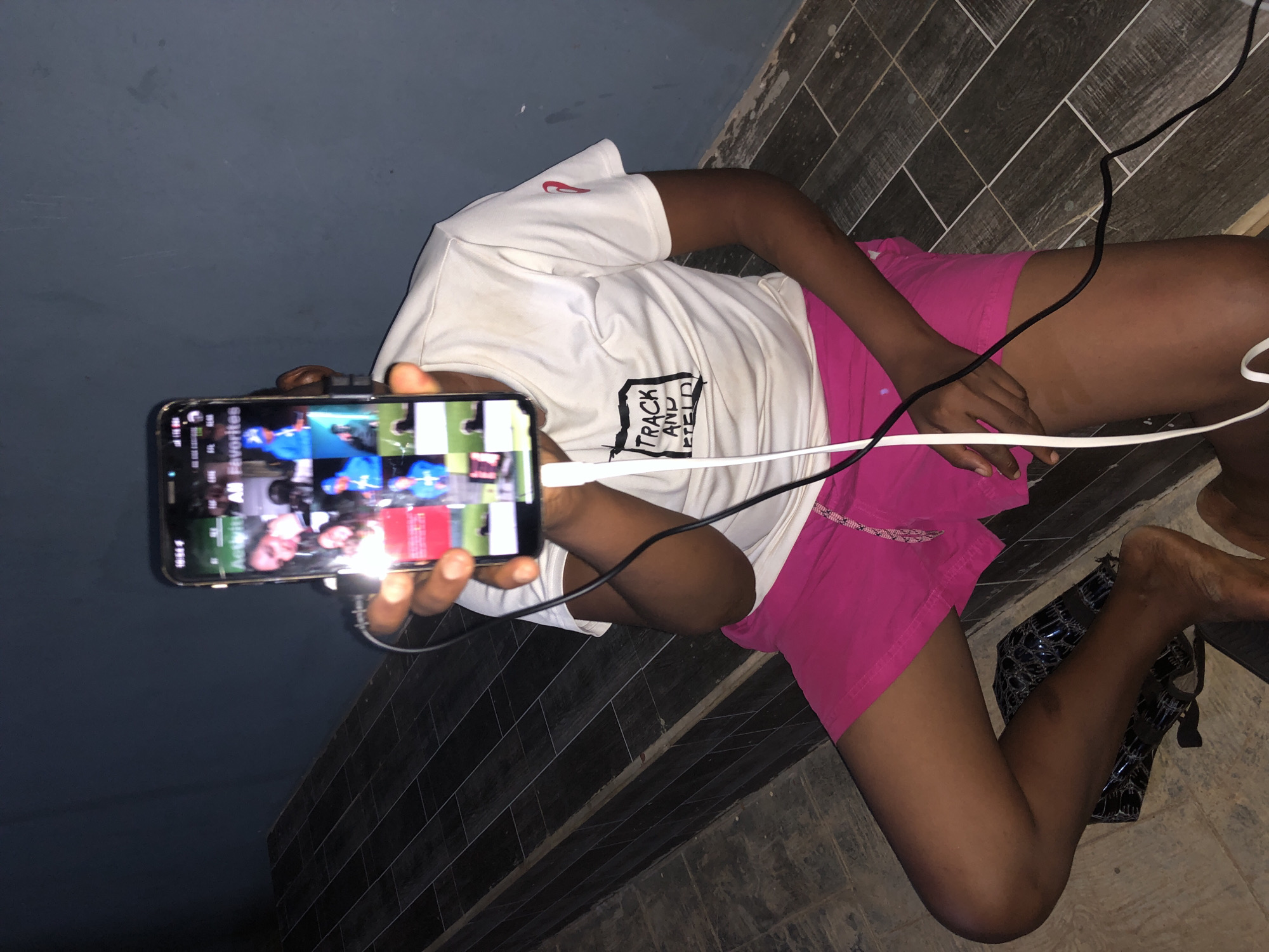 A person is lying on a gray tiled floor with a dark gray wall in the background. The person is wearing a white t-shirt with "TRACK AND FIELD" printed on it and pink shorts. They are holding a smartphone in their hand, displaying a video or images on the screen. Two charging cables, one white and one black, are plugged into the phone and extend across the person's body. Next to the person's foot is a pair of black sandals with a patterned texture.