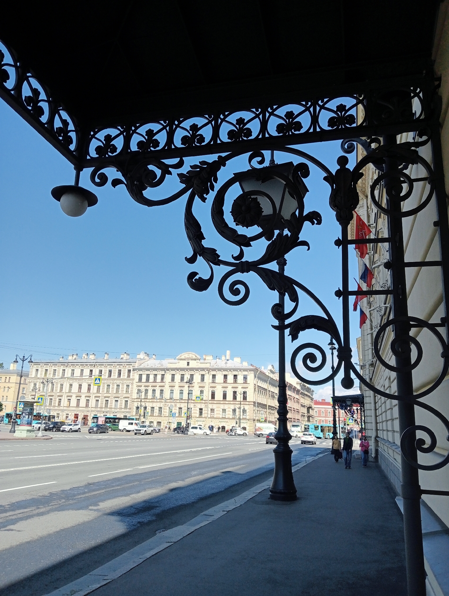 In Saint Petersburg, Russia, a detailed shot captures a street scene on a clear, sunny day. The foreground features an ornate, dark wrought-iron canopy with decorative scrolling and floral motifs, casting shadows on the pavement. A lamppost, also made of dark metal, extends upwards from the sidewalk, its intricate design mirroring the canopy. The sidewalk is made of grey concrete, and a wide asphalt road runs parallel to it, with visible white lane markings. Several cars, including a white van, a white SUV, and a dark-colored sedan, are driving on the road. Pedestrians, two women and a man, are walking on the sidewalk in the middle ground. One woman wears a pink jacket and blue jeans, another wears a yellow jacket and dark pants, and the man is dressed in a dark jacket and blue jeans.
In the background, a long, multi-story building with a light yellow facade dominates the skyline. The building features numerous windows and decorative architectural elements, including a row of flags displayed on its facade. The sky above is a bright, cloudless blue. Further down the street, more buildings and traffic are visible, suggesting a bustling urban environment. The overall impression is one of a well-preserved historical city, with classical architecture and a lively streetscape.