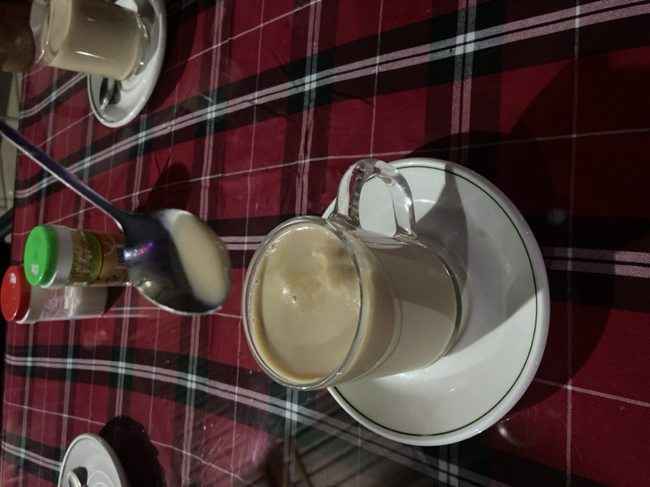 The image presents a high-angle, slightly oblique view of a table setting, likely in an indoor establishment in Bagamoyo, Tanzania. The table is covered with a vibrant red, black, and white plaid tablecloth.
The primary subject is a clear glass mug, filled with a light brown, frothy beverage that appears to be milky tea or coffee. This mug rests on a white ceramic saucer with a thin green rim. To the left of the mug, a large, reflective silver spoon lies on the tablecloth, holding a small pool of the same light brown liquid in its bowl, suggesting it has just been used for stirring or tasting the drink.
Adjacent to the spoon are two small, translucent plastic shakers or bottles, possibly containing spices or condiments. One has a bright green cap, and the other a red cap. Some faint, unreadable text or a label is visible on the green-capped shaker.
In the upper left corner of the frame, another white saucer with a similar beverage cup is partially visible, indicating multiple servings. A small portion of a silver can, possibly a soda can, can also be seen at the bottom left edge of the image.
The lighting appears subdued, casting soft shadows from the glass mug and the spoon onto the tablecloth, which suggests an evening setting or ambient indoor lighting. The scene conveys a relaxed atmosphere, perhaps a break time or a casual meal where beverages are being enjoyed. No people are visible.