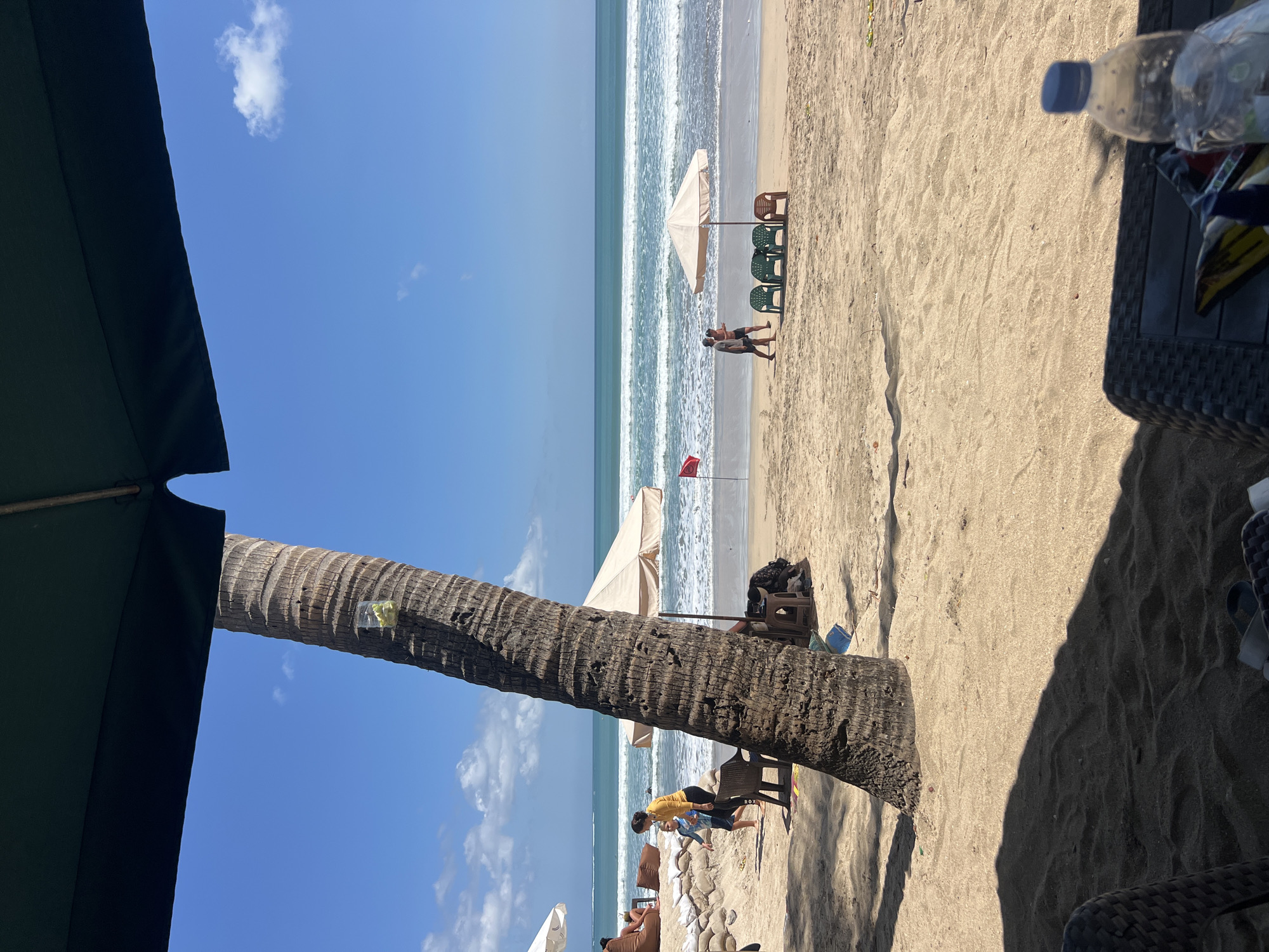 The image depicts a sun-drenched beach scene in Kuta, Indonesia. The foreground is dominated by the textured trunk of a palm tree, casting a shadow across the sandy ground. Partially visible in the lower right corner is outdoor furniture, including a dark-colored table with a water bottle on it.
The middle ground features a sandy beach with a few scattered beach chairs and umbrellas. A beige umbrella stands over a cluster of green and brown plastic chairs. Further back, the turquoise ocean meets the shore with gentle waves. In the background, the horizon is marked by the vast blue sky dotted with a few white clouds.
Several people are present on the beach. A woman in a yellow top and dark leggings walks with a young boy in a blue and white patterned shirt. Further down the beach, two individuals are walking towards the ocean. Some people are seated under umbrellas or on beanbag chairs in the distance. A red flag is planted in the sand near one of the umbrellas. The overall impression is that of a typical, lively day at a popular beach destination.