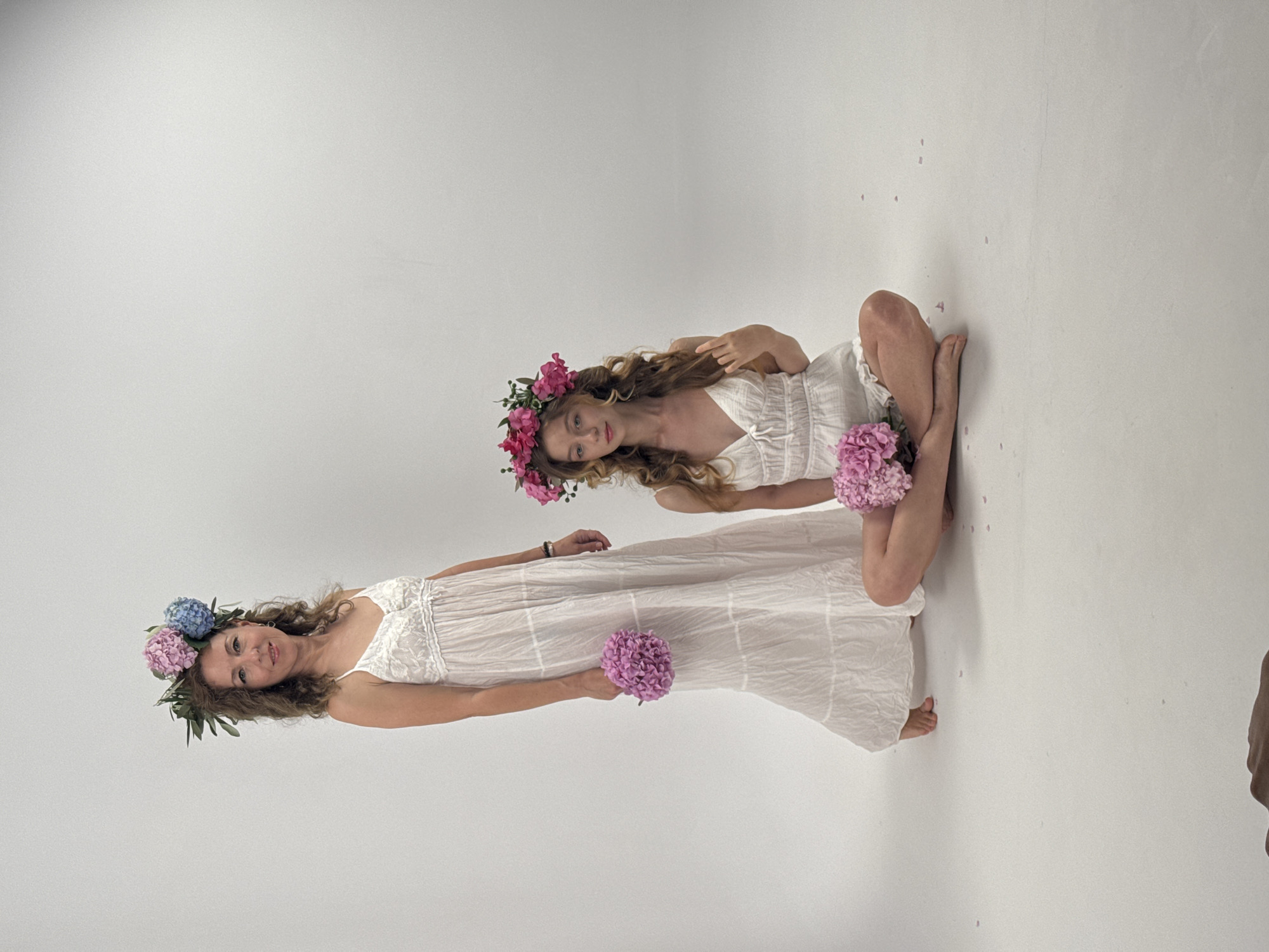 Two women are posing for a photograph in a studio with a plain white background. The woman on the left is standing and wearing a long, white, tiered dress with a floral crown made of pink and blue flowers. She is holding a bouquet of pink flowers in her right hand. The woman on the right is sitting on the floor, wearing a white romper with a floral crown made of pink flowers. She is holding a bouquet of pink flowers in her lap. Petals are scattered on the floor around her. The image does not contain any identifiable landmarks or indications of Budva, Montenegro.