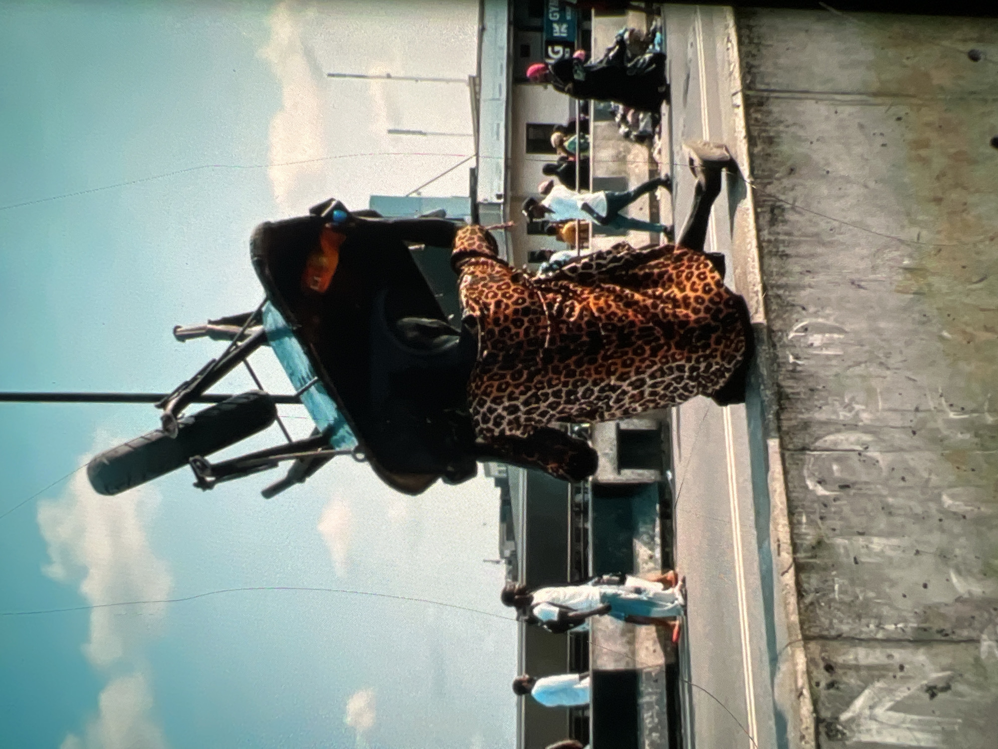 A person wearing a leopard-print garment carries a large object resembling a wheelbarrow on their head, balancing it with outstretched arms. The individual is positioned on a wall, with their right leg extended forward. In the background, several other people are visible on a street, some walking and others standing. Buildings and a cloudy sky are also part of the environment. The overall scene suggests a busy urban setting, likely in Lagos, Nigeria, given the context provided.