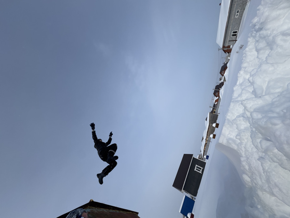 This image depicts a dynamic outdoor scene in Gornyi (Dvurechenskoe S/P), Russia, under a clear, light blue sky. The environment is heavily covered in snow, indicative of winter conditions. In the upper left quadrant, a single individual, dressed in dark winter attire including a jacket, pants, and gloves, is captured mid-air, suspended against the sky. Their body is oriented horizontally, with arms outstretched and legs bent, suggesting a jump or fall. The left arm is slightly higher than the right, and the left leg is more acutely bent.
The foreground consists of a large, uneven accumulation of bright white snow, forming a substantial snowdrift or pile. Below the airborne individual and extending into the midground, several residential structures are visible. A prominent, dark-colored house with a brown sloped roof and a single white-framed window is situated on a snow-covered incline. To its left, a partial blue structure is discernible. Further into the background, a cluster of additional houses with snow-covered roofs and varying construction materials (including grey block and darker, possibly wooden or brick, facades) can be seen. Utility poles with overhead lines are present in the distance. The overall perspective is a low-angle shot, with the horizon line notably tilted upwards from left to right, emphasizing the height and motion of the airborne person. Daylight illuminates the scene uniformly.