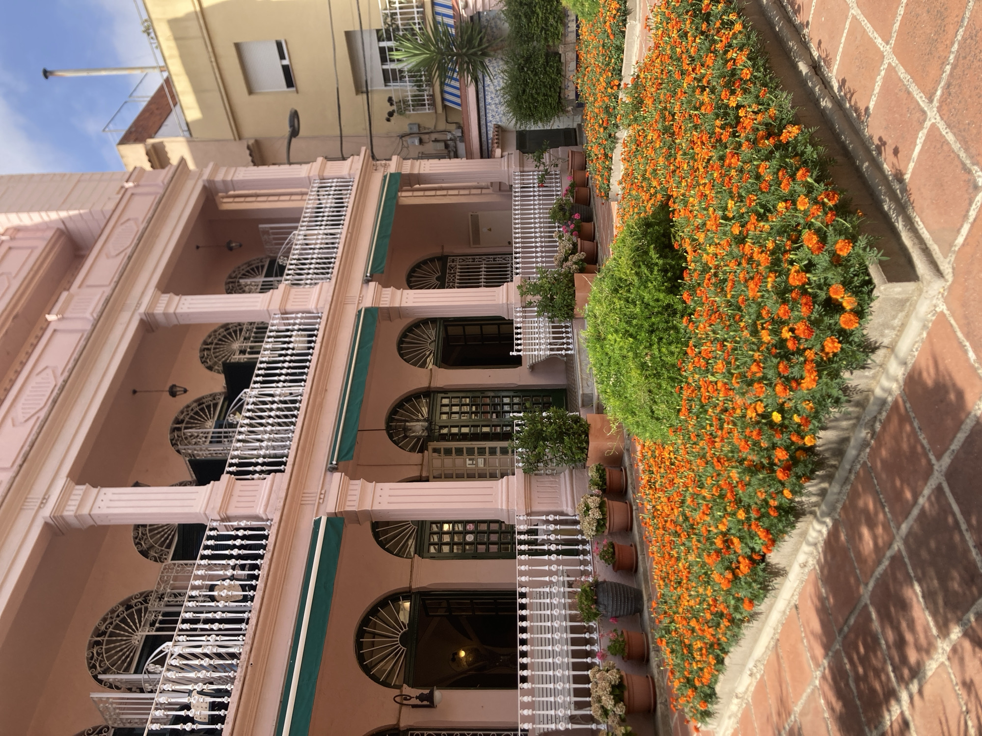 This is a full-shot, eye-level, outdoor photograph taken in Lloret de Mar, Spain. The image features a light pink, multi-story building with an arched balcony and ornate white railings. In the foreground, a rectangular garden bed is densely planted with bright orange marigold flowers, bordered by a low concrete wall and a red tiled patio. The building has green awnings over arched doorways and windows. Adjacent to the pink building, a yellow building with a white-framed window is visible. The sky is partly cloudy.