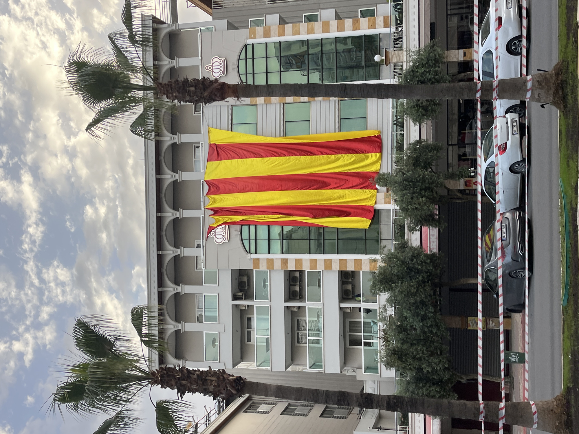 The image captures a street scene in Alanya, Turkey. In the foreground are two palm trees, one on each side of the frame. Behind them is a multi-story building with a large Catalan flag draped across its facade. The building appears to have a hotel or commercial function, indicated by its architectural style and the presence of what appear to be air conditioning units. To the right, cars are parked along the street, and a striped barrier is partially visible. The sky is cloudy.