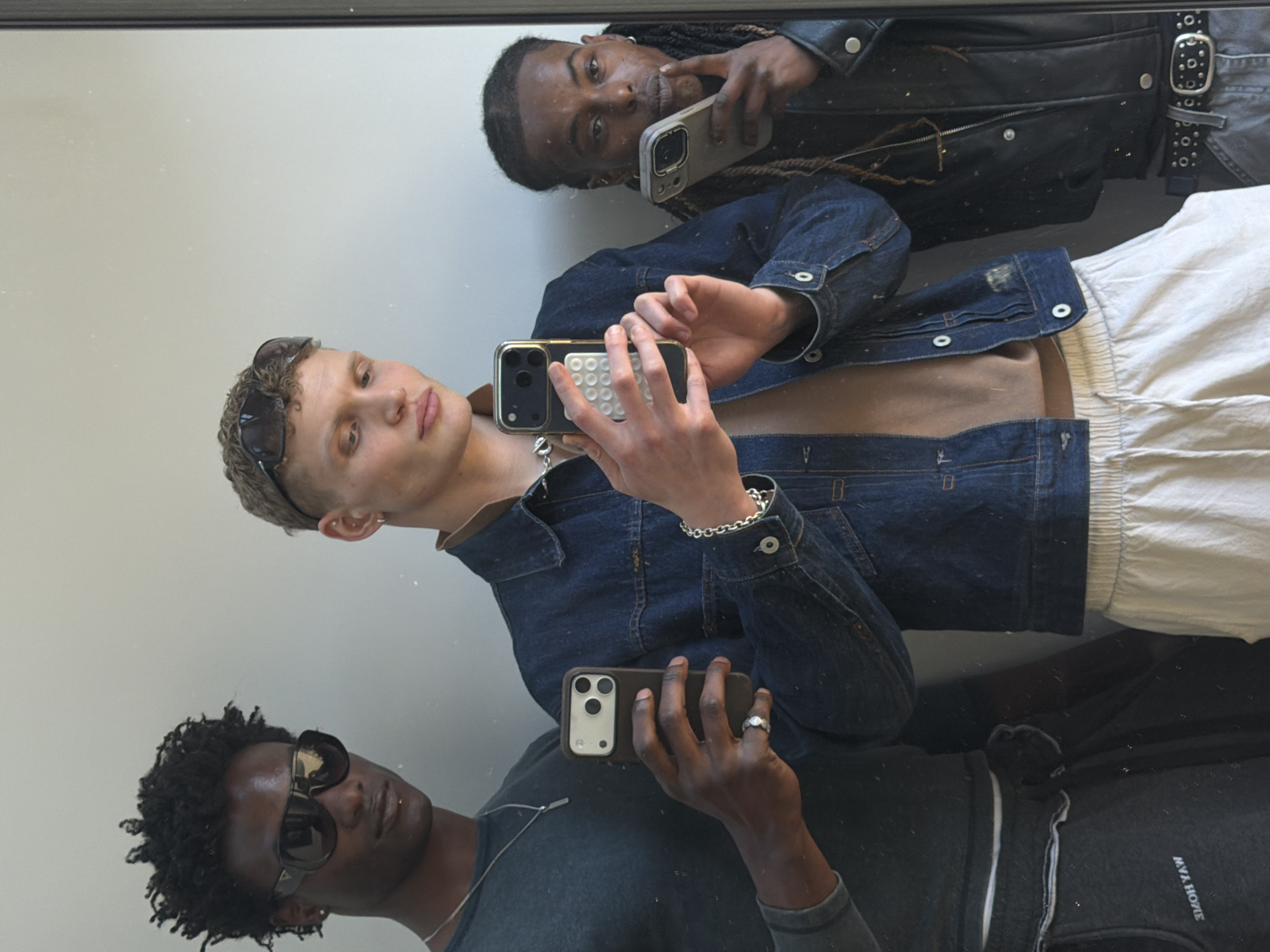 The image is a mirror selfie featuring three young men in an indoor setting with a plain, light-colored wall reflected behind them. The photo is rotated 90 degrees counter-clockwise from a standard portrait orientation.
The central figure is a light-skinned man with short, curly blonde hair, wearing sunglasses on his head. He is dressed in a denim jacket over a brown t-shirt, light-colored pants, a silver chain, and a silver link bracelet. He holds a dark smartphone with a white pop-socket-like attachment.
To his right (top of the image), a dark-skinned man with dreadlocks is visible. He wears a black leather jacket, a dark belt with a silver buckle, and holds a light-colored smartphone. He is posed with one hand near his chin.
To his left (bottom of the image), another dark-skinned man with short, dark curly hair and dark sunglasses is shown. He is wearing a dark t-shirt, a silver chain, and a ring on his left hand. He holds a dark smartphone. The reversed text "MVA HOME" (likely "MYA HOME" when un-mirrored) is visible on his dark grey pants or shorts.
All three men are actively taking a mirror selfie with their smartphones. The expressions are generally neutral and posed. The mirror surface appears to have some dust or minor imperfections. While the context of London, United Kingdom, is provided, there are no specific location cues visible within the image itself.
