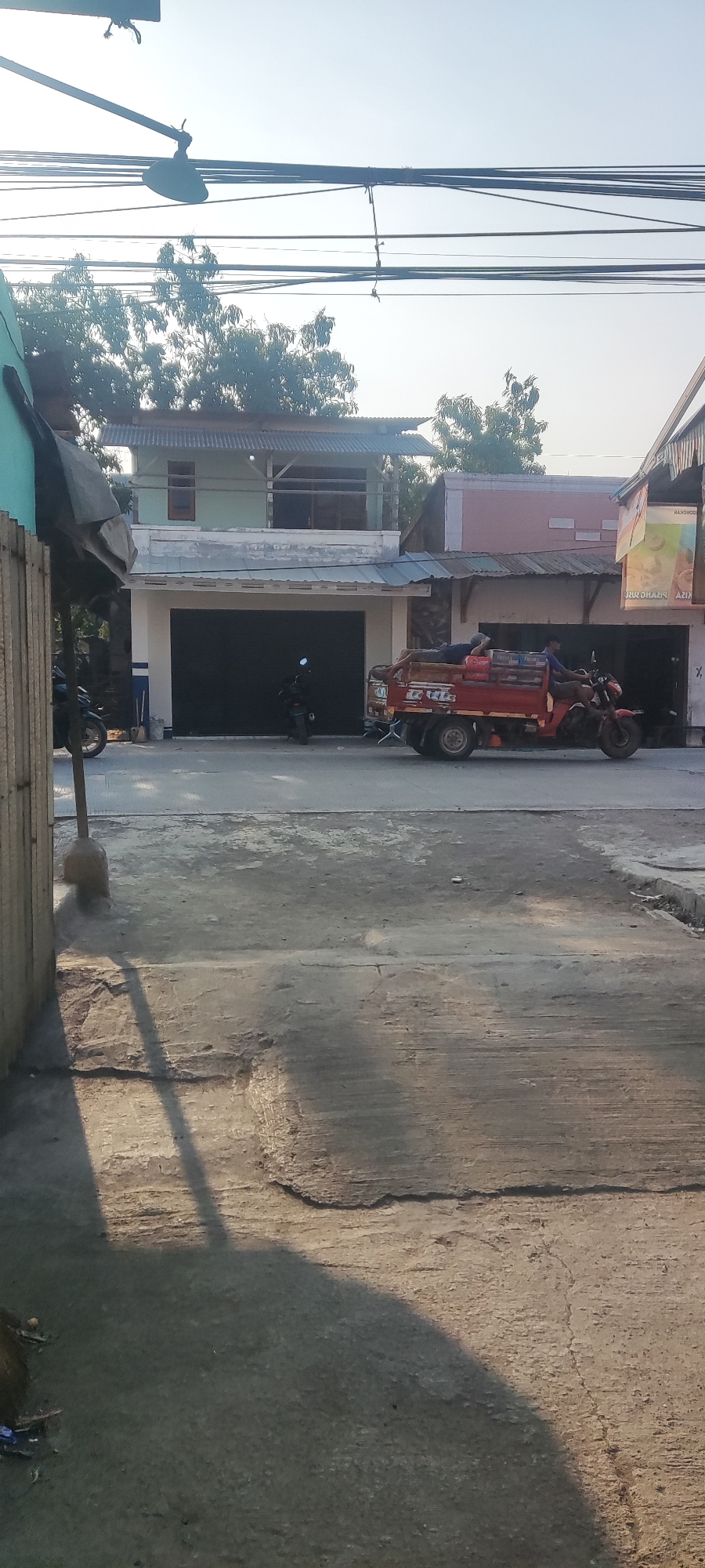 The image depicts a street scene in Gebang, Indonesia. In the foreground, a concrete surface shows signs of wear and tear, with cracks and rough textures. A long, dark shadow stretches across this surface, cast by an unseen object to the left.
In the middle ground, a red three-wheeled truck is parked on the street. Two individuals appear to be resting on top of the truck's cargo, which is covered with various items, including what look like crates or boxes. The truck has a utility bed and is positioned in front of a building with large, dark garage doors.
To the left of the truck, a black motorcycle is parked. A second motorcycle, also black, is visible further down the street.
The background features a multi-story building with a corrugated metal roof. Trees with dense foliage are visible behind the building. Overhead, a network of power lines crisscrosses the sky. The lighting suggests it is either early morning or late afternoon, with the sun casting long shadows. The overall environment appears to be an urban or semi-urban setting in Indonesia.