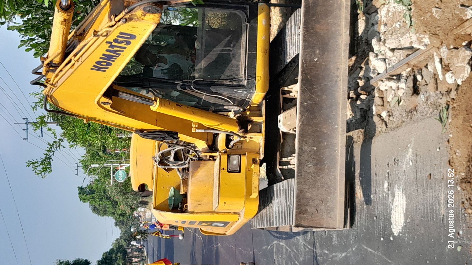 An excavator is parked on a road under construction in Takeran, Indonesia. The excavator is yellow and has "KOMATSU" written on its side. The excavator's blade is lowered to the ground, and there are piles of dirt and debris around it. The road is partially paved, with some areas showing exposed concrete. In the background, there are trees, utility poles, and a clear blue sky. A few people can be seen in the distance, presumably working on the construction site. A timestamp in the lower left corner reads "21 Agustus 2026 13.52".
