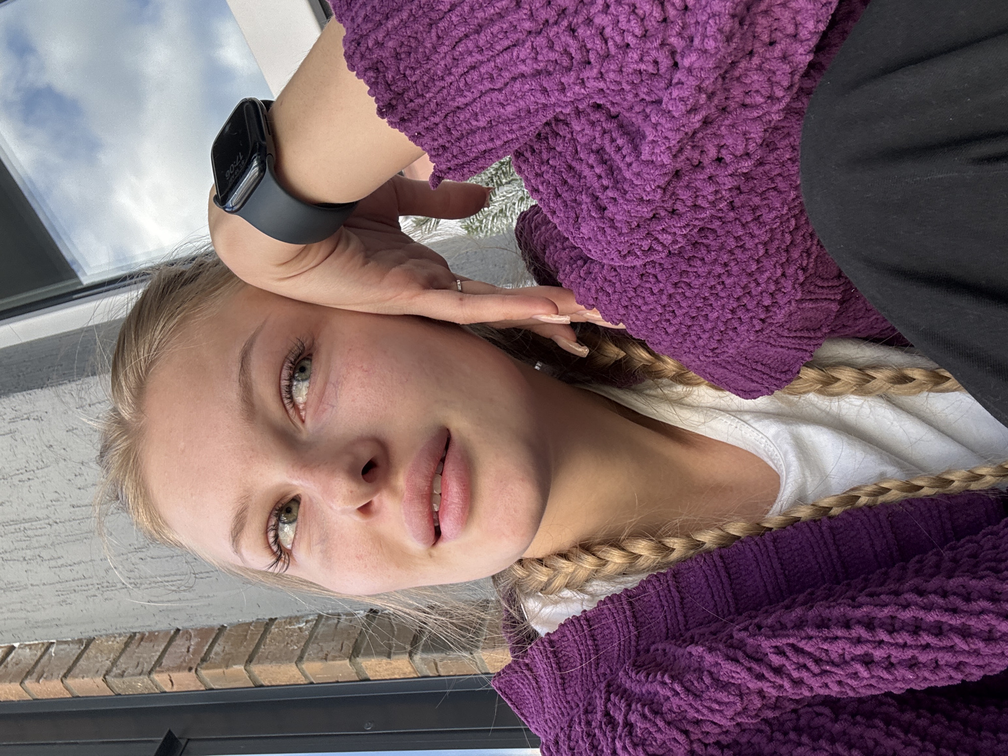The image shows a close-up of a young woman with blonde hair in braids, wearing a purple knitted sweater over a white t-shirt. She is looking to her left, with her left hand resting on her cheek. She is wearing a smartwatch on her left wrist. The background is blurred, but appears to be an outdoor setting with brick and a window reflecting the sky. The lighting suggests it is daytime. There are no specific landmarks or details visible that would allow for the identification of the city of Perm, Russia.