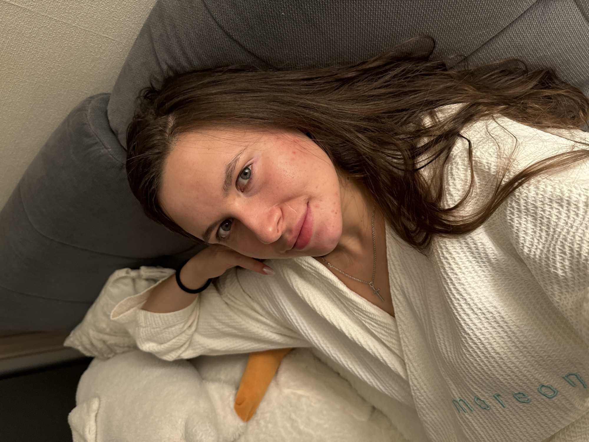 A young woman with long, dark brown hair is captured in a close-up, high-angle shot, reclining on a grey upholstered surface, likely a sofa or bed. She is wearing a white, textured robe, possibly with a waffle weave, which features the word "moreon" embroidered in light blue on the sleeve. A silver chain with a small cross pendant adorns her neck, and a black hair tie is visible on her left wrist.
The woman looks directly at the camera with a calm and relaxed expression, offering a soft smile. Her head is gently resting on her left hand. Her eyes are green or hazel, and her complexion shows some reddish blemishes. Below her left arm and the white robe, part of a fluffy white object, possibly a plush toy or pillow, and an orange element (like a beak or limb) are visible, contributing to a cozy and intimate indoor setting. The background consists of the solid grey upholstery. The image conveys a sense of comfort and repose in what appears to be a private residence or accommodation in Akulovo (Gp Kubinka), Russia.