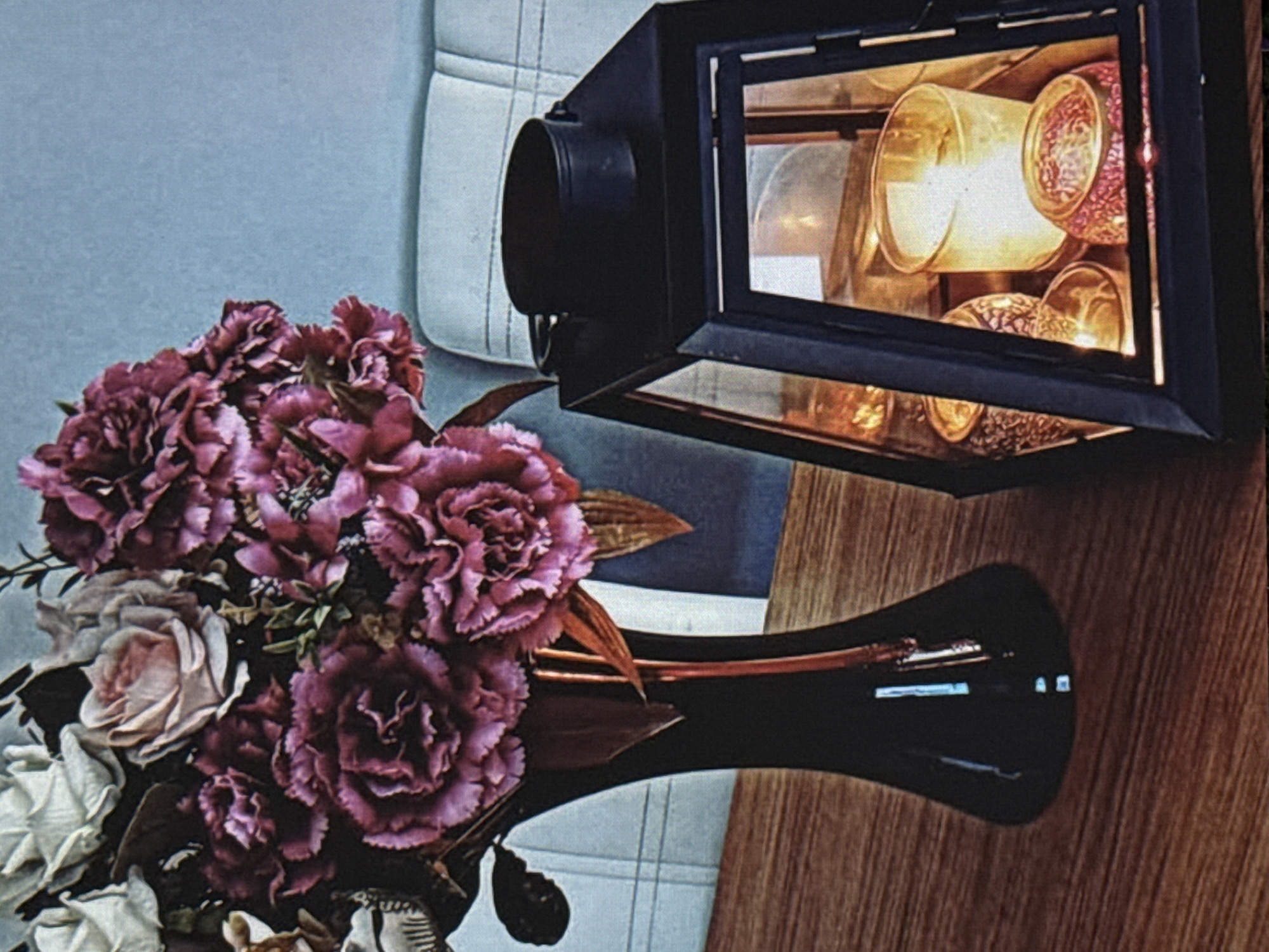 This media file captures a still life arrangement featuring a bouquet of artificial flowers and a decorative lantern. The bouquet, predominantly in shades of deep pink and burgundy with some lighter pink and white accents, is partially submerged in a glossy black vase. The flowers appear to be carnations or similar multi-petaled blooms, with some delicate greenery interspersed. The vase itself has a sleek, modern shape.
To the right of the flowers and vase is a black lantern with glass panels. Inside the lantern, several glass candle holders are visible, illuminated from within by a warm, golden light, suggesting candles are lit or the lantern is powered by an artificial light source. The candle holders have textured glass, adding visual interest.
The background consists of two distinct elements. To the left, there's a textured surface that appears to be light blue or gray, possibly a wall or upholstery with a subtle pattern. To the right and behind the lantern, a wooden surface with a prominent grain pattern is visible, suggesting a table or countertop. The overall composition is framed by the edges of these elements and the dark tones of the lantern and vase. The lighting suggests an indoor setting, possibly in the evening or a dimly lit room, given the illuminated lantern. There is no visible text in the image.