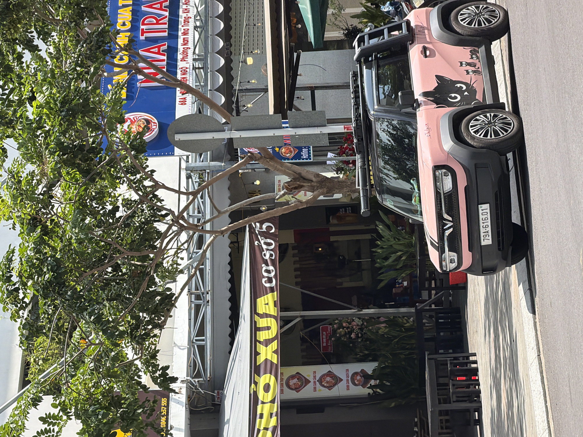 In Nha Trang, Vietnam, a pink electric car adorned with black cat decals is parked on a street. The vehicle has a roof rack and a ladder at the back. To the left of the car, there is a restaurant with outdoor seating and a banner displaying the text "PHỞ XUA cơ sở 5" and other Vietnamese phrases. Behind the restaurant, a large blue sign with Vietnamese text, including "NHATRANG," is visible. The street is paved, with a sidewalk in front of the restaurant. Trees with green foliage are present, casting shadows on the ground. The overall scene suggests a typical urban environment with commercial establishments and a parked vehicle.