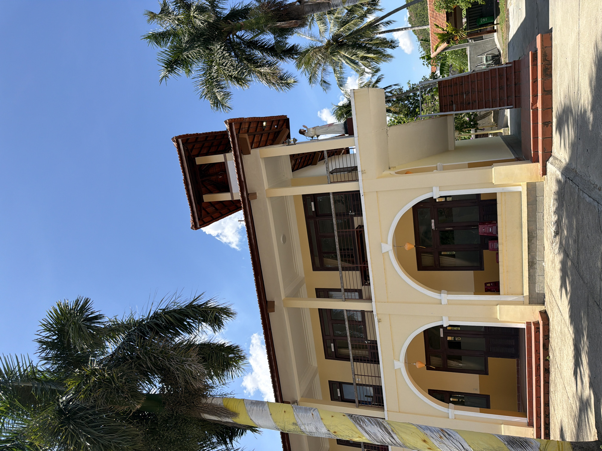 This image captures a sunny day in Hội Tín, Vietnam, featuring a multi-story building with a yellow facade and a red-tiled roof. The building has several balconies and arched entrances on the lower level, with dark-framed windows and doors. A statue of a standing figure, possibly religious, is visible on an upper balcony. Tall palm trees frame the scene, with one prominently featuring yellow and white striped decoration on its trunk. Red brick stairs lead to an external staircase on the side of the building. The ground is paved, and shadows from trees are cast across it, suggesting a tropical environment. The sky is a clear blue with scattered white clouds.