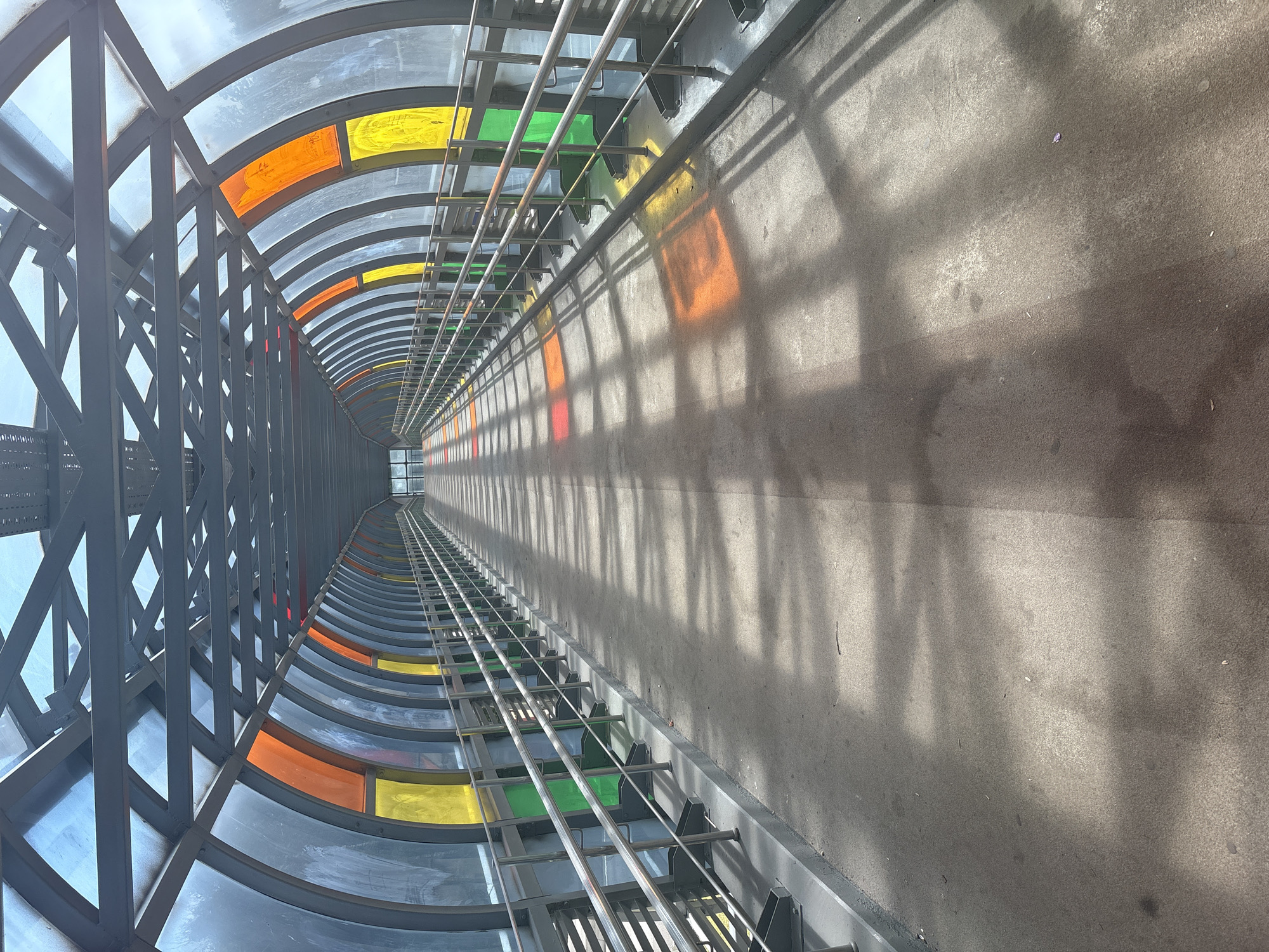 The image depicts a pedestrian overpass with a curved, covered walkway. The walkway has a metal framework supporting translucent panels on its sides and roof. These panels are interspersed with sections of colored glass in green, yellow, and orange. The walkway floor appears to be concrete, and a metal railing runs along both sides. Sunlight streams through the colored panels, casting patterned shadows on the walkway. The scene is devoid of any people or animals. The overpass is situated in the city of Vatutinki, Russia.