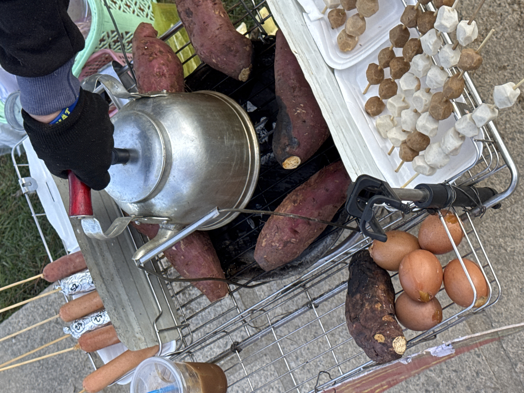 A high-angle shot reveals a food vendor's metal cart in Da Lat, Vietnam. A gloved hand steadies a silver cooking pot atop a grill filled with roasted sweet potatoes. To the right, skewers of fish balls rest on white trays, while a basket holds boiled eggs and another sweet potato. Skewered sausages are also visible. The backdrop includes pavement and sparse vegetation.