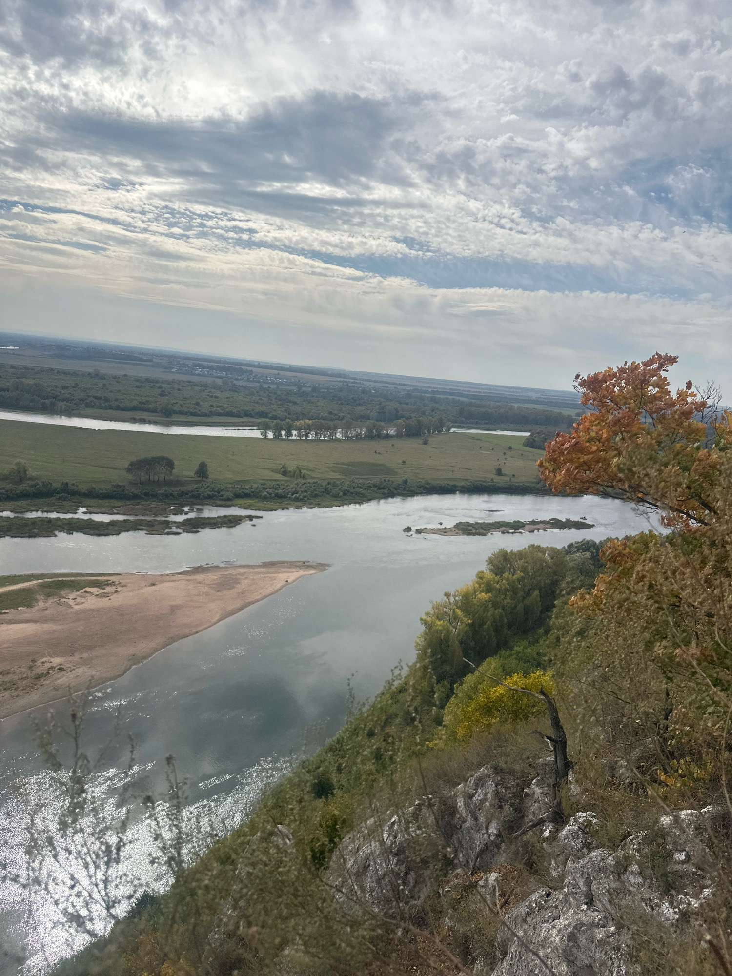 The image captures a panoramic view from a rocky, elevated vantage point overlooking a wide river and a fertile plain in Simskii, Russia. The sky is filled with a dynamic pattern of white and gray clouds, hinting at an overcast or partly cloudy day. In the foreground, the rocky terrain is covered with sparse vegetation, including dry grasses and some autumn-colored foliage on bushes and trees.
A large river dominates the middle ground, with a sandy embankment on the left side and islands of vegetation in the water. The river reflects the sky and sunlight, creating bright, shimmering patches on its surface. The plain on the opposite side of the river is a patchwork of green fields and scattered trees, with a line of trees marking the distant horizon. Small settlements or buildings are faintly visible in the far distance, nestled amongst the trees.
The overall scene conveys a sense of natural beauty and vastness, with the river carving its path through the landscape and the surrounding environment showcasing the transition into autumn. There are no people or animals visible in the image.