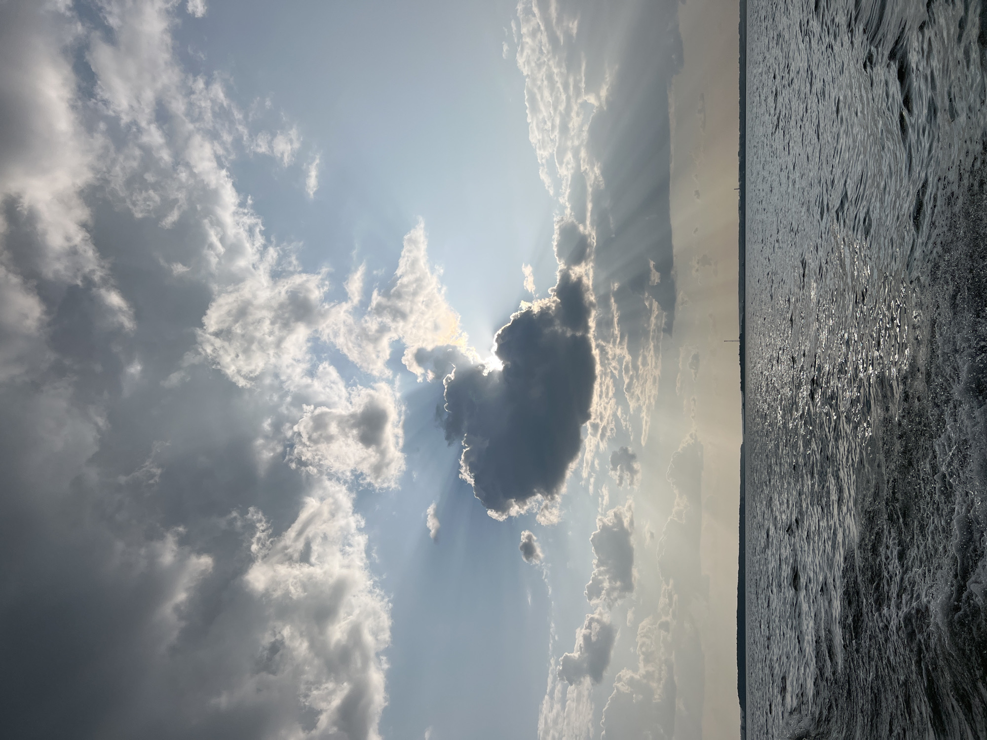 The image shows a wide shot of a body of water with a dark, choppy surface. In the background, a distant, flat shoreline is visible under a dramatic sky filled with clouds. The sun is partially obscured by a large, dark cloud, casting visible rays of light through breaks in the clouds. The sky is a mix of bright blue and dark gray clouds, suggesting a dynamic weather condition. The foreground shows the white wake of a boat moving through the water. The overall scene conveys a sense of movement and natural drama. The location is identified as the city of Novopichugovo, Russia. There are no people or specific objects in the scene, other than the implied presence of a boat creating the wake.