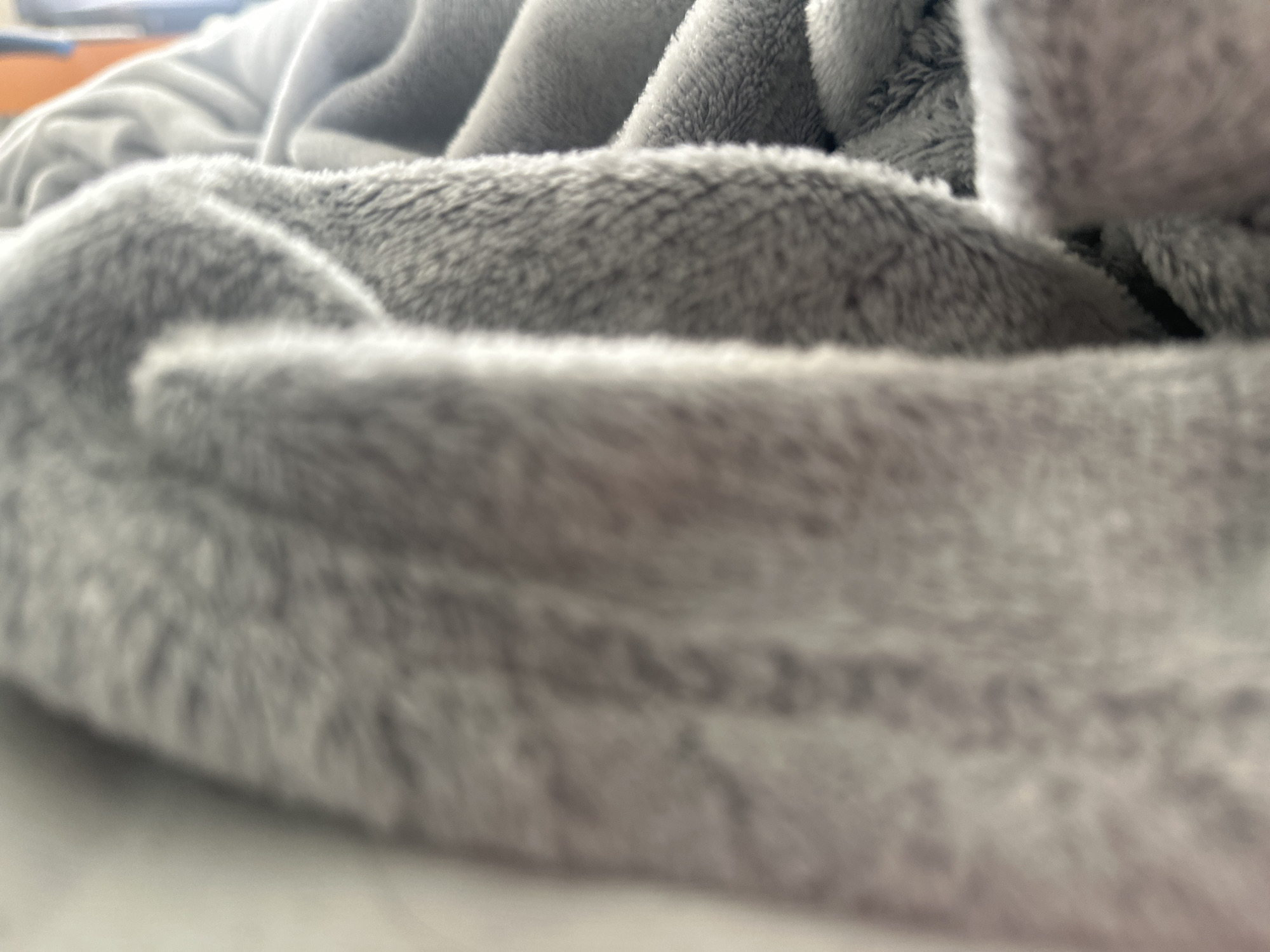 The image shows a close-up view of a gray, fuzzy blanket. The blanket is soft and appears to be a plush material. The lighting is soft and diffused, highlighting the texture of the fabric. There are no people, animals, or other objects visible in the image. The environment is indoors, with a hint of an orange object in the upper left corner that may be a piece of furniture or clothing. The scene is static and peaceful. There is no information available about the location in the city of Shumen, Bulgaria, as this is an abstract image of a blanket.