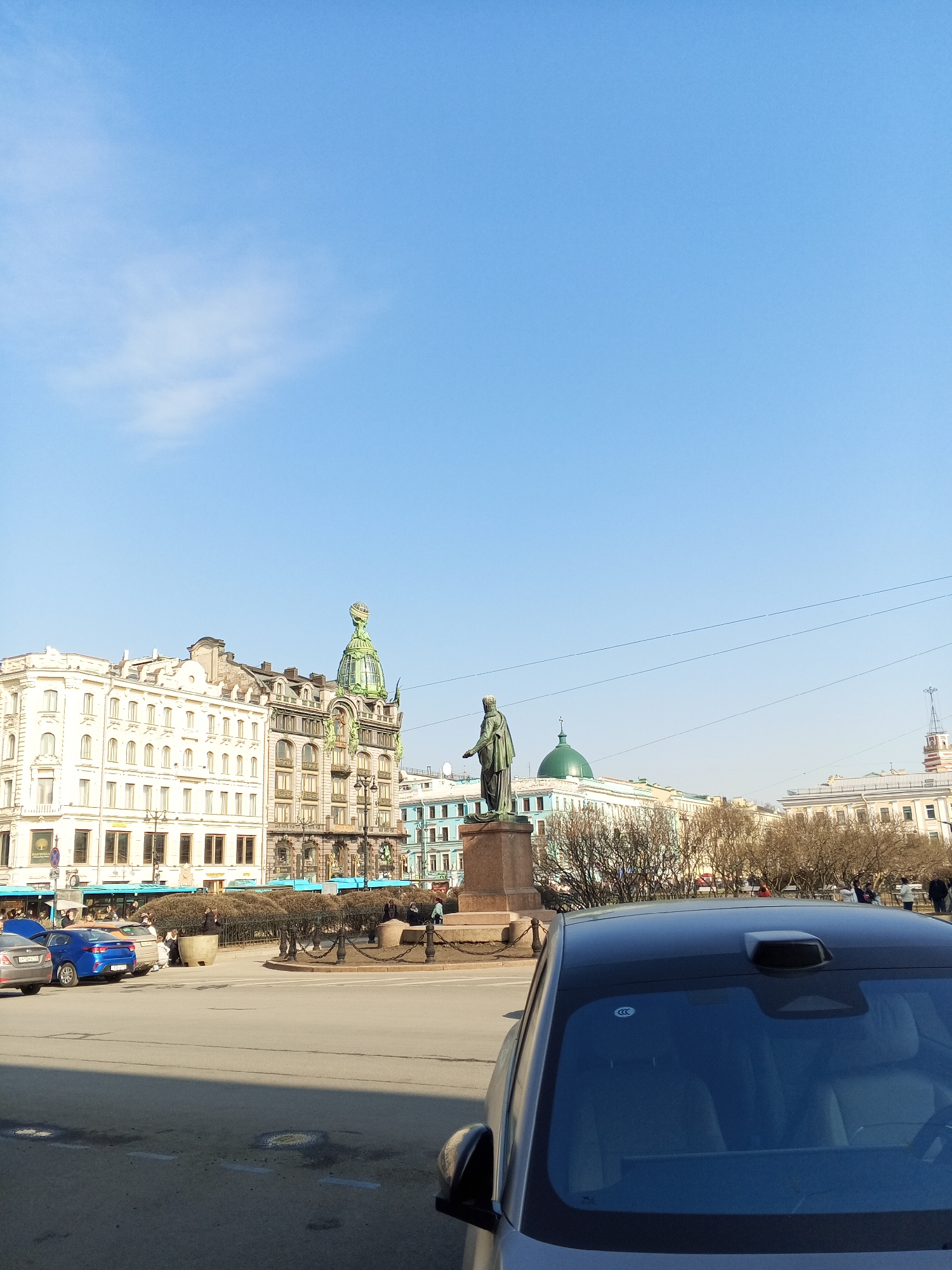 The image shows a cityscape in Saint Petersburg, Russia, featuring Anichkov Bridge and the monument to Catherine the Great. The foreground contains the rear and side of a car, parked on what appears to be a paved street. Behind the car, a plaza is visible, with a statue of Catherine the Great centered on a pedestal. The statue faces left. Buildings are prominent in the background, including the Singer House, with its distinctive glass dome. To the right of the statue, another building with a green dome is visible. The sky is a clear blue with a few scattered clouds. The atmosphere suggests a bright, sunny day. A few people are scattered around the plaza.