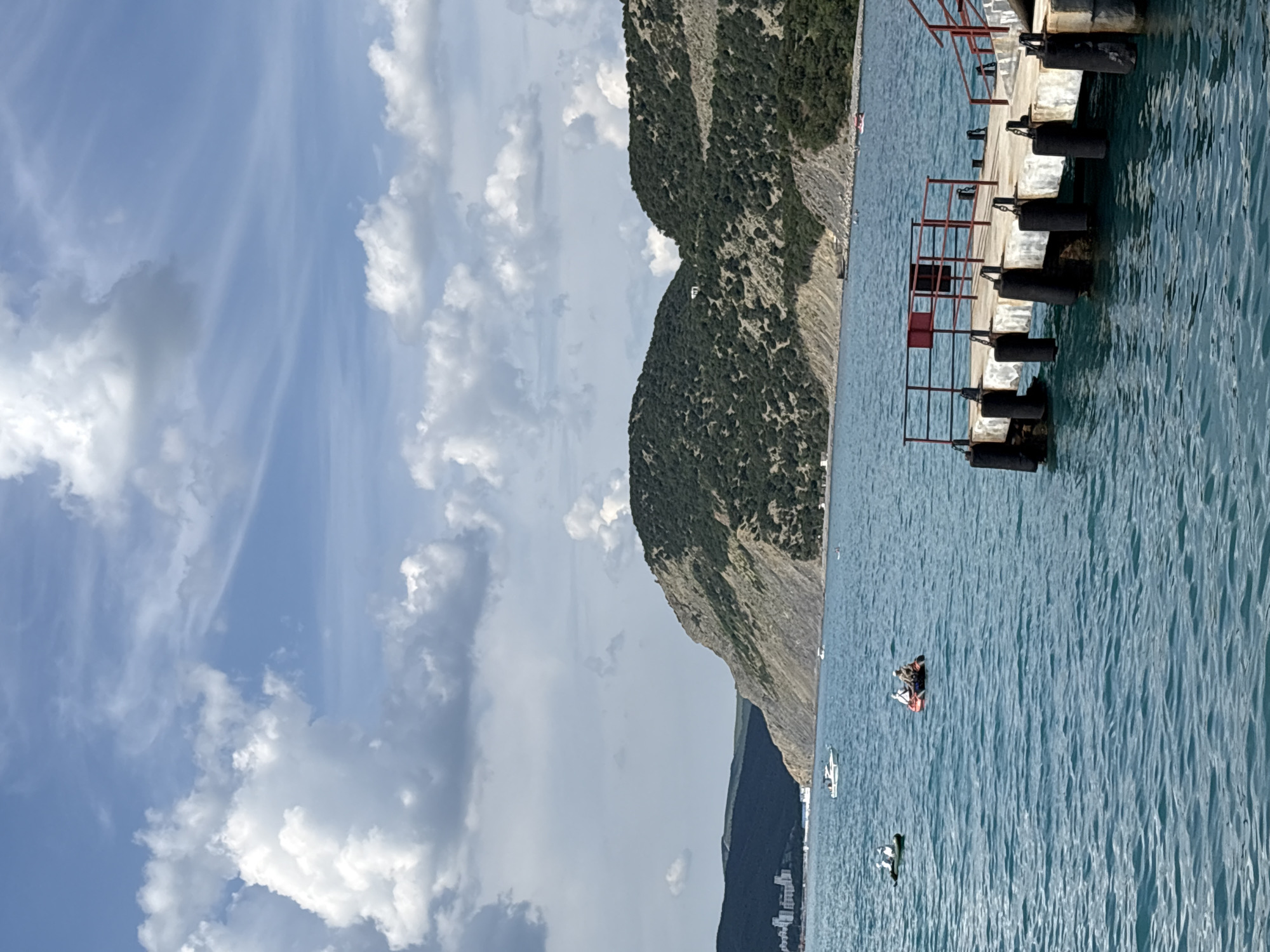 This image captures a coastal scene in Bol'shoi Utrish, Russia. A concrete pier with dark red and black metal railings extends from the lower right into calm, turquoise-blue water. Black cylindrical fenders are attached along the pier's side. In the mid-ground, an orange-red inflatable dinghy with two individuals, one wearing a light-colored hat, is positioned on the water. To its left, a greenish-white kayak or small boat carries a single person. A larger white motor yacht is visible further back in the water, along with several other small boats closer to the distant shore. The background is dominated by steep, rugged mountains covered in dense dark green vegetation, rising directly from the sea. Visible rock strata are present on the mountain slopes. On the far left, a cluster of white buildings is situated at the base of the mountains along the coastline. The sky above is partly cloudy, featuring large white cumulus clouds and thinner cirrus clouds against a light blue backdrop.