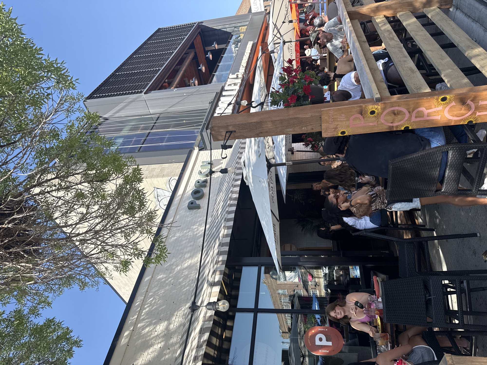 This image depicts an outdoor dining scene at a restaurant called "Porch" located in Calgary, Canada. The restaurant has a modern architectural style with a brick facade and a multi-story glass and metal extension. People are seated at tables on a patio, enjoying food and drinks. Several umbrellas provide shade. A wooden sign with "Welcome to The Porch" and sunflower motifs is visible. The sky is clear and blue, suggesting a sunny day. The general atmosphere is casual and social.
