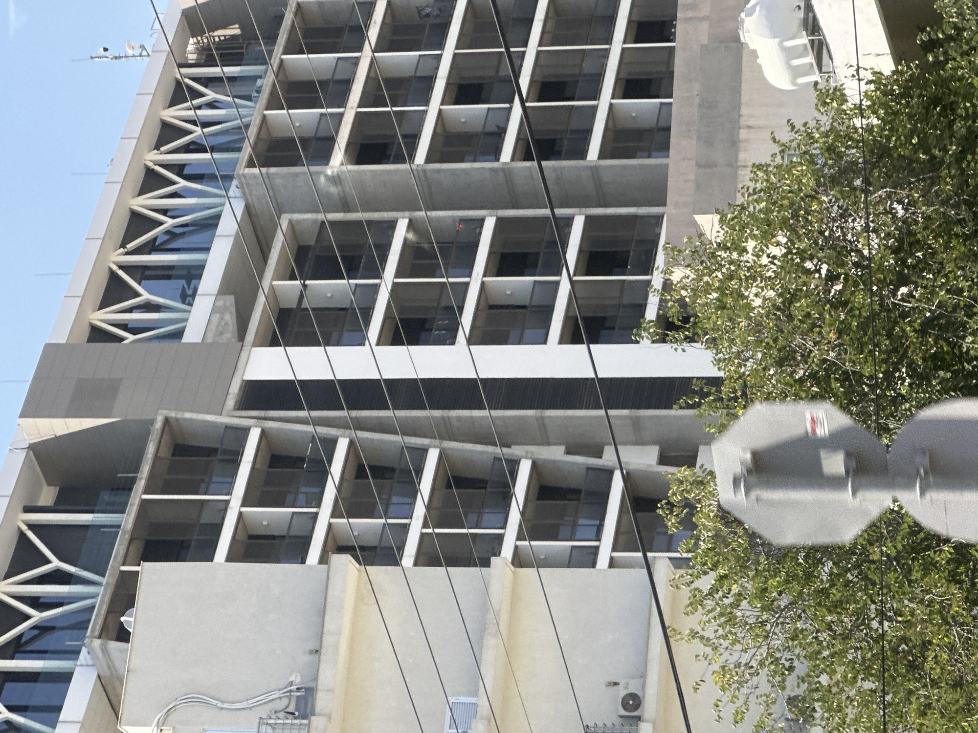 The image shows a modern building with many windows and balconies. The building is tall and appears to be made of concrete and glass. There are some trees and branches in the foreground, and power lines are visible crisscrossing the frame. A white water tank is visible on the roof of a lower section of the building. A gray octagonal traffic sign is also partially visible in the lower center of the image. The sky is clear and blue.