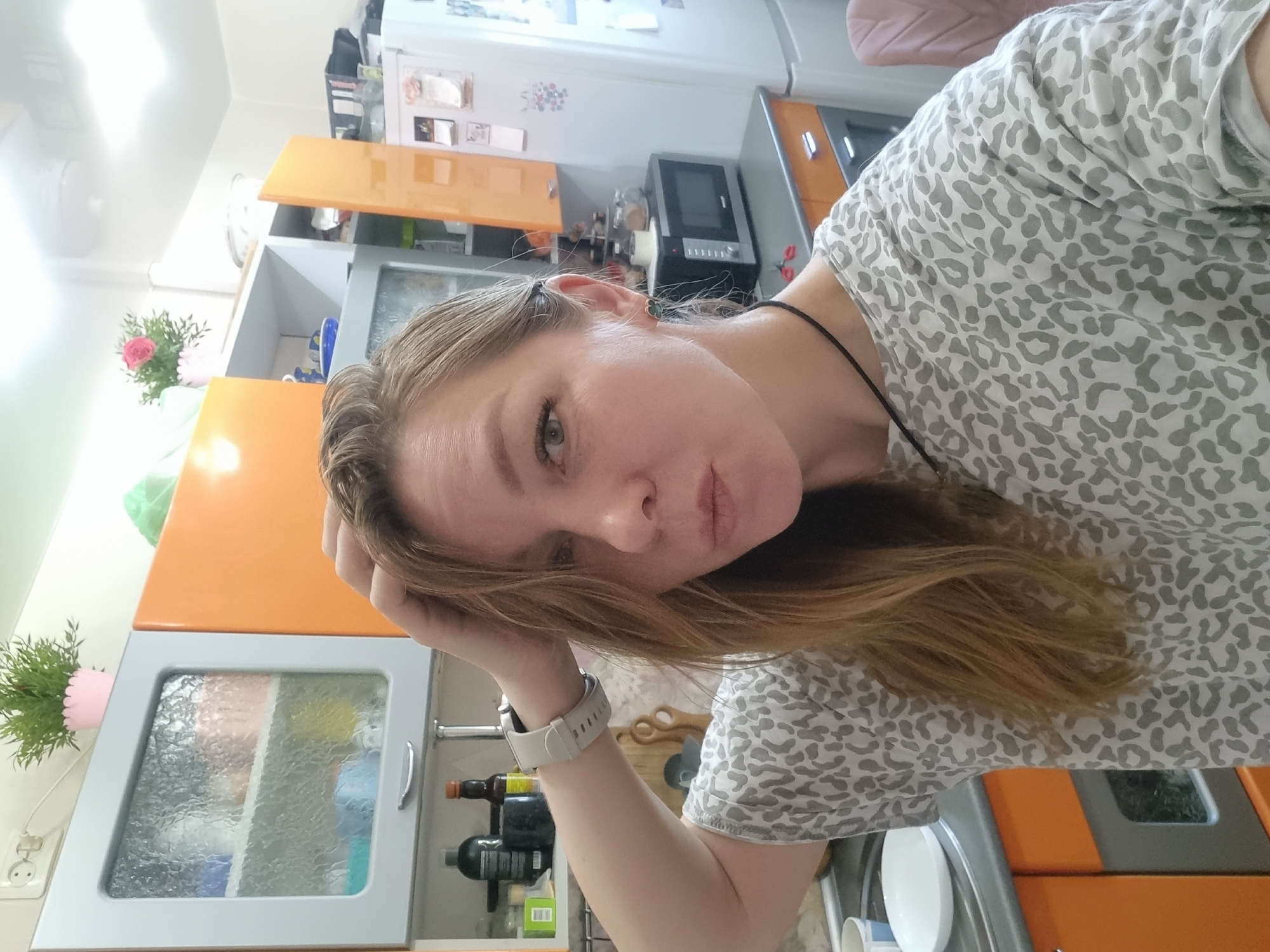 This image is a selfie of a woman taken in a kitchen in Chaikovsky, Russia. The photo is oriented 90 degrees counter-clockwise. The woman, with long, light brown hair, is positioned in the foreground, looking directly at the camera with a neutral to slightly pouting expression. Her left hand is casually placed on her head or hair. She is wearing a light-colored t-shirt with a grey leopard print pattern and a thin black string around her neck. A white smart watch is visible on her left wrist.
The background reveals a functional kitchen space with bright orange and grey/silver cabinets. Some lower cabinets feature frosted glass doors, through which various kitchen items are faintly visible. A microwave oven and a large white refrigerator are also present. The refrigerator door appears to have magnets or small pictures attached. To the left, a green potted plant in a pink pot is visible, and another potted plant with pink flowers is on a shelf. The lighting is bright, suggesting daytime conditions indoors. There is a power outlet on the wall near one of the plants. No discernible text is visible on any objects or surfaces.