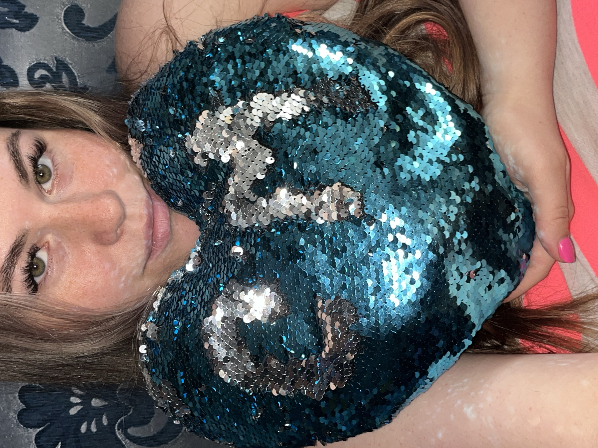 This image, captured in Chaikovsky, Russia, presents a close-up, top-down view of a person, likely female, lying down and holding a heart-shaped sequin pillow. The subject's left eye, a striking green or hazel, is visible, framed by prominent eyelashes. Her long, flowing brown hair is spread around her head and shoulders. Several distinct light white patches are visible on her skin, particularly on her left cheek, forehead, and extending onto her visible arm, suggesting a skin condition.
The central object is a large, vibrant heart-shaped pillow covered in reversible sequins. The dominant color of the sequins is a deep teal or blue, which can be flipped to reveal a bright silver. Intricate patterns have been formed on the pillow by manipulating these sequins, creating silver designs against the blue background.
The person's partially visible lips suggest a soft, relaxed expression. A glimpse of her hand shows fingers with bright pink nail polish, resting near the pillow. The background features various fabrics: a dark blue material with a lighter, ornate floral or paisley pattern on the left, and a solid reddish-pink surface, likely a bed or sofa, on the right. The scene depicts an intimate indoor setting, conveying a sense of calm and comfort. No visible text is present.