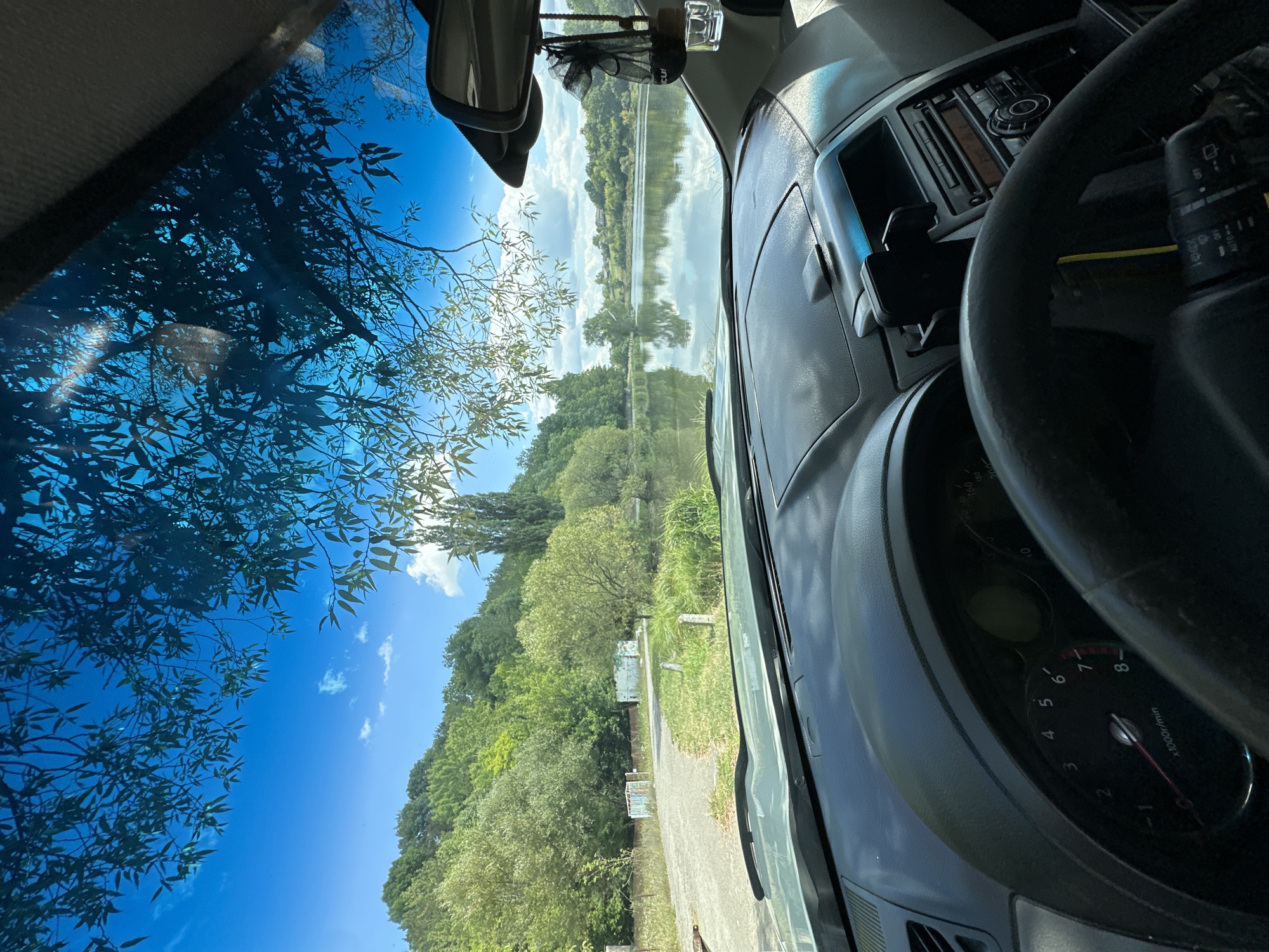 The image is a photograph taken from inside a car, looking out through the windshield at a scenic view of a lake and surrounding hills. The interior of the car is visible in the foreground, including the dashboard, steering wheel, and a rearview mirror. The exterior scene features a calm lake reflecting the blue sky and fluffy white clouds. Dense green trees and a hillside cover the background. The location is identified as Makiv, Ukraine.