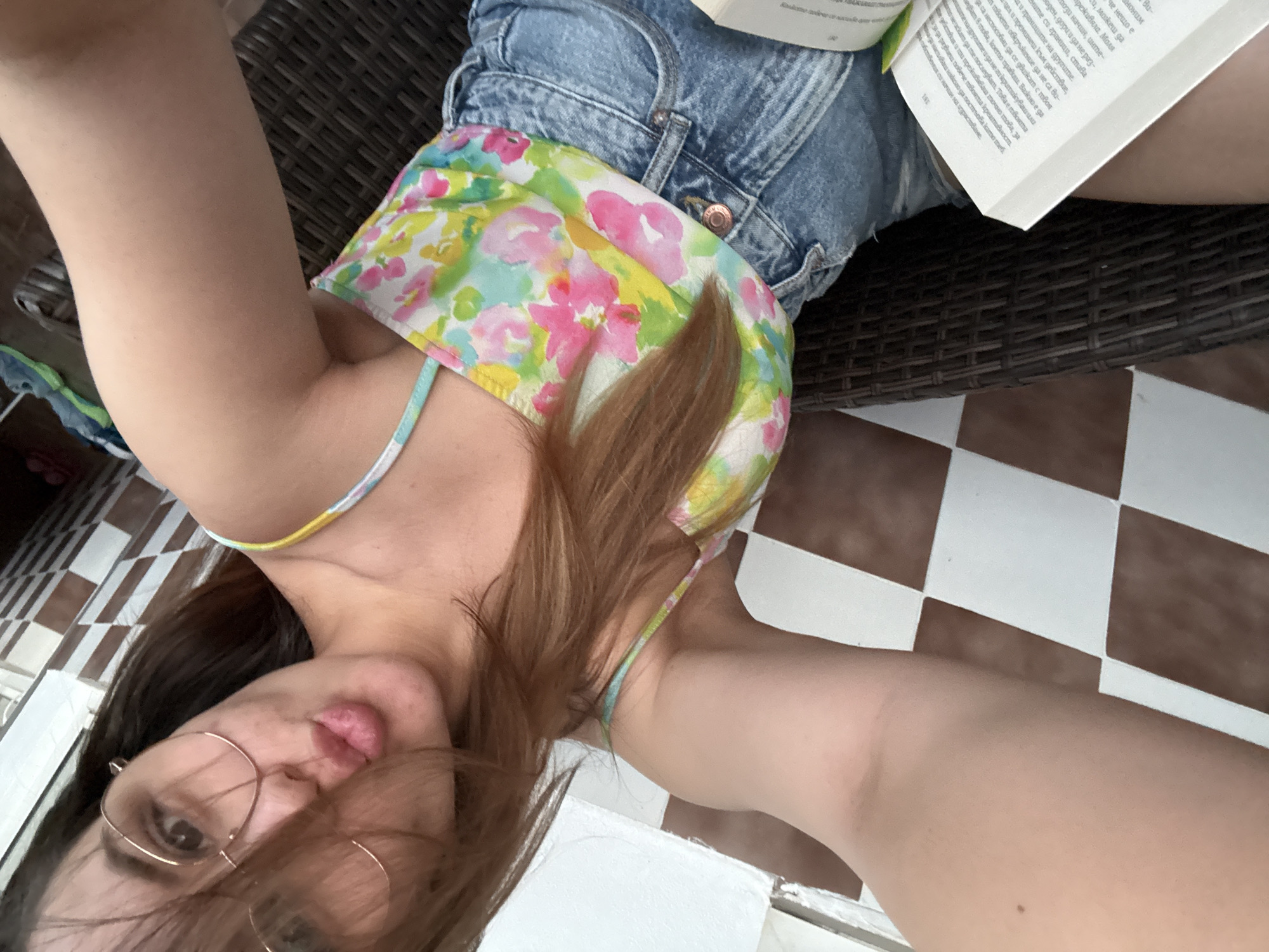 A woman with glasses and brown hair is taking a selfie while sitting and reading a book. She is wearing a floral top and denim shorts. The environment includes a checkered tile floor and wicker furniture. Text in Bulgarian is visible on the open book, suggesting the location might be in Bulgaria, potentially Sofia.