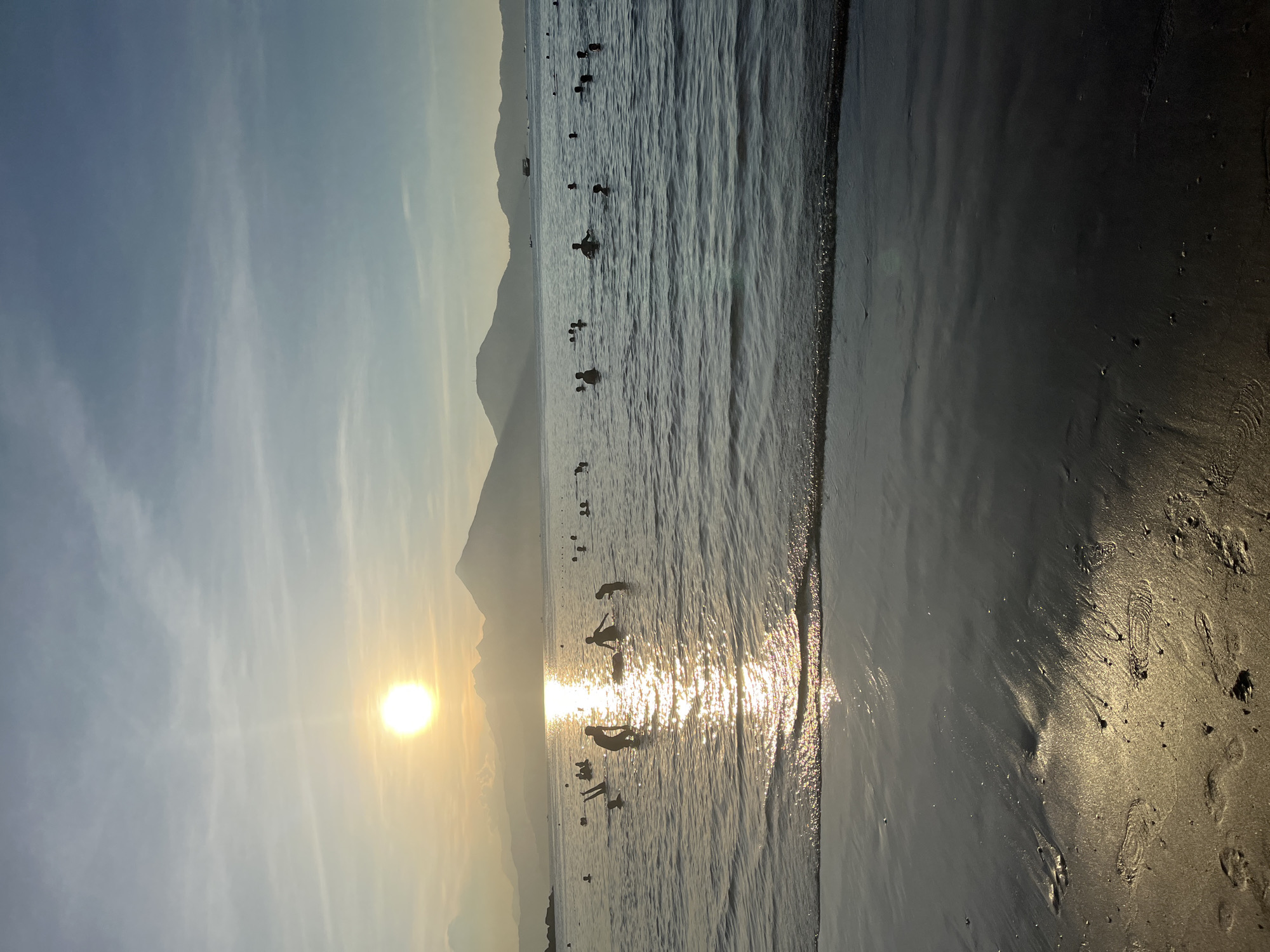 This image depicts a tranquil coastal scene during either sunrise or sunset in Đà Nẵng, Vietnam. The sun, a bright golden disc, is low on the horizon, casting a brilliant, shimmering reflection across the water's surface that extends towards the foreground.
Silhouetted against the bright reflection and the sky are numerous individuals in the water, appearing to be swimming, wading, or simply enjoying the sea. Their forms are distinct dark figures against the sparkling water, suggesting a leisure activity. In the background, a range of large mountains rises, their distant peaks appearing as dark silhouettes against the softer sky, defining the coastal landscape.
The sky exhibits a gradient of colors, transitioning from a light blue at the top to warmer golden and orange hues closer to the sun. Wispy, delicate clouds are scattered across the sky, catching the soft light. The foreground reveals a stretch of wet beach sand, appearing dark and reflective, particularly where it catches the sun's golden glow. Some footprints are visible on the sand, indicating human presence.
The overall impression is one of peace and natural beauty, with the warm light of the low sun creating a picturesque and contemplative atmosphere. There is no visible text in the image.