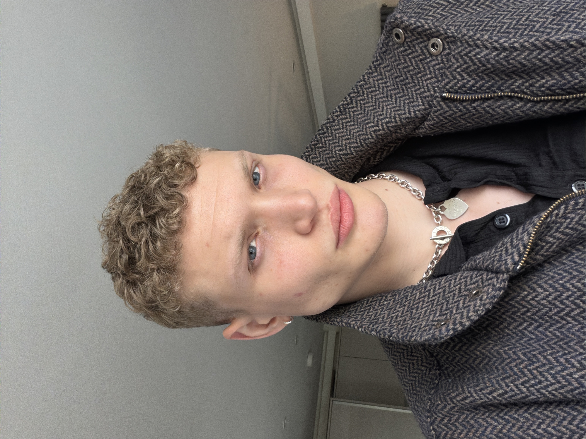 A close-up portrait, oriented sideways, features a young man with light, curly, blondish-brown hair and distinct blue eyes. He gazes directly at the camera with a neutral expression. He is wearing a dark, possibly black, textured shirt beneath a jacket with a prominent dark and light herringbone pattern. The jacket features metallic zippers and snap buttons. Around his neck, a chunky silver chain necklace is visible, from which hangs a heart-shaped pendant. Visible text on the pendant clearly reads "TIFFANY & CO.". A small silver hoop earring is present in his left ear. The background consists of a plain, light grey wall, indicating an indoor setting. Hints of a light-colored structure or furniture are visible on the right side of the frame. The lighting is even, illuminating the subject's face and upper body. The context provided indicates the location is London, United Kingdom.