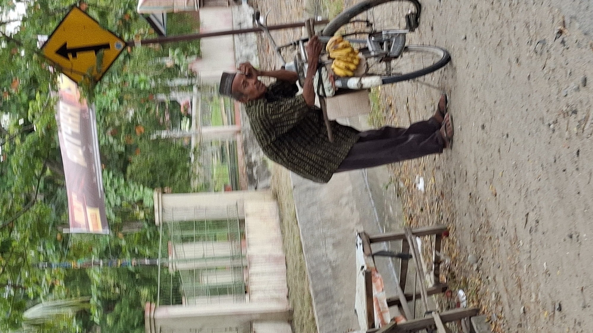 The image depicts a man standing next to a bicycle with a bunch of bananas attached to it. He is wearing a dark patterned shirt, dark pants, and a cap. The man is leaning on the handlebars of the bicycle with his head resting on his hand, appearing to be tired or contemplative. The bicycle has a basket in front where the bananas are placed.
In the background, there is a concrete barrier, a fence, and some trees. A yellow diamond-shaped road sign with a black arrow pointing upwards is visible on the right side of the image, indicating a directional change or a lane merge. A banner with some text is hanging above the trees. The ground is covered with dirt and scattered leaves.
The scene appears to be a roadside, possibly in Lhokseumawe, Indonesia, given the context provided. The overall atmosphere is casual and somewhat rural.