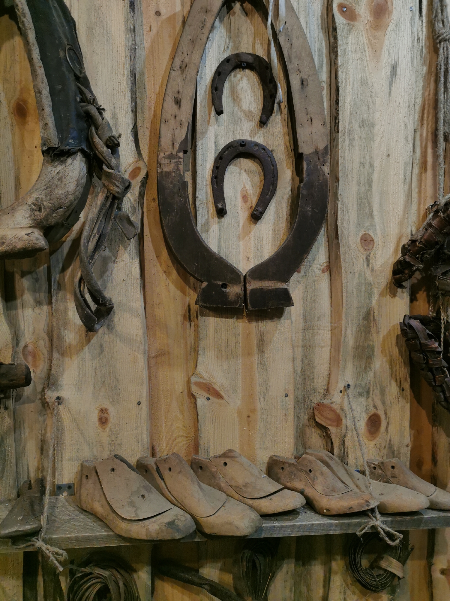 This is a medium shot of a collection of antique blacksmithing and shoemaking tools displayed on a wooden wall. Dominating the upper portion of the image are two large, rusty metal forms that appear to be part of a horseshoe or a similar forging tool, with two smaller horseshoes hanging from them. To the left, a weathered leather boot and some metal straps are attached to the wall. The lower part of the image features a wooden shelf holding several wooden shoe lasts, which are molds used for making shoes. The wall itself is made of rough, light-colored wooden planks, with visible knots and grain. The lighting is dim, casting shadows and highlighting the textures of the aged objects. The scene evokes a rustic, workshop atmosphere. The location is in Шчарчоўскі сельскі Савет, Belarus.