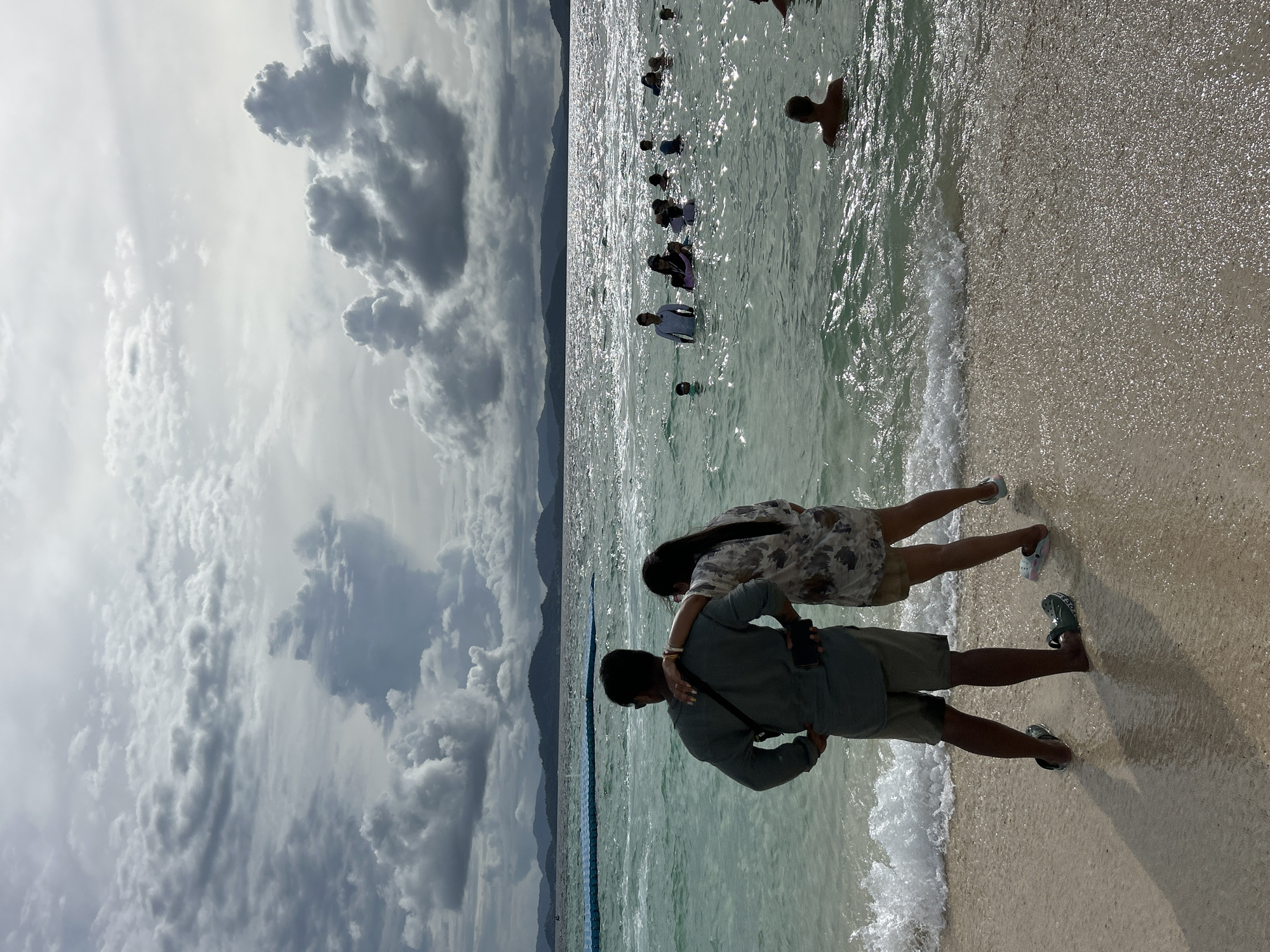 In a scenic Thai beach setting, several people are enjoying the water and shore. The day appears bright with sunlight reflecting off the water. Dark, dramatic clouds dominate the sky, suggesting a dynamic weather system. In the foreground, a couple stands on the sandy beach, with the man embracing the woman from behind. They are both looking out towards the ocean. The woman wears a floral-patterned dress, and the man is dressed in a green shirt and khaki shorts. The sea is calm with gentle waves lapping at the shore. Further out in the water, a group of people are wading and swimming, their figures silhouetted against the bright sunlight. The image captures a moment of leisure and relaxation at a tropical beach.