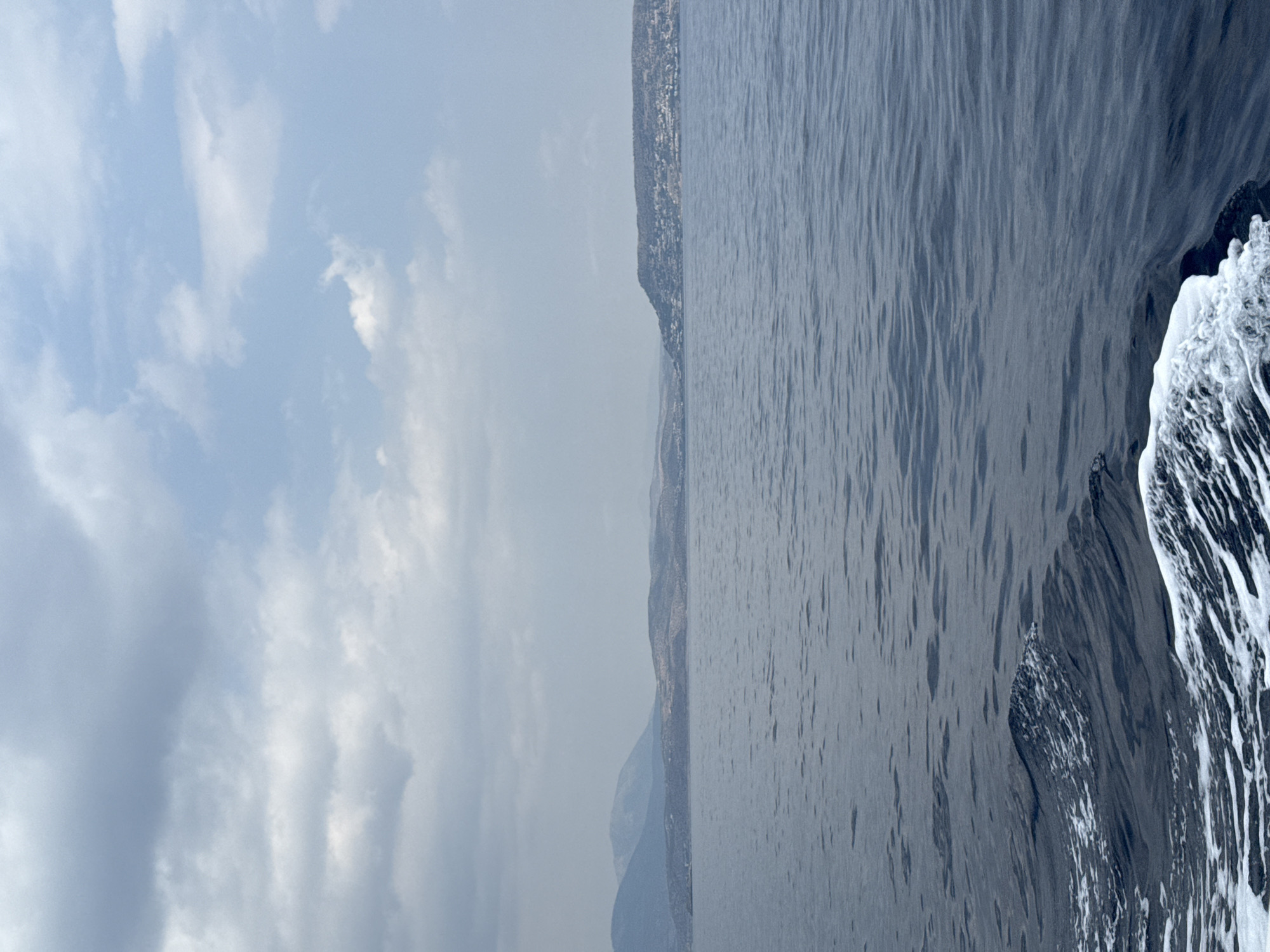 The image depicts a view of the sea from a moving boat. The water is dark blue and rippled, with white foam trailing from the boat's wake in the foreground. In the distance, a hazy coastline is visible with rolling hills and some scattered buildings. The sky above is overcast with grey and white clouds, suggesting a diffused light. The scene captures a sense of travel and a coastal landscape. It is likely taken near Saronida Municipal Unit, Greece, given the characteristic coastal topography and vegetation. There are no people or specific animals visible in the frame.