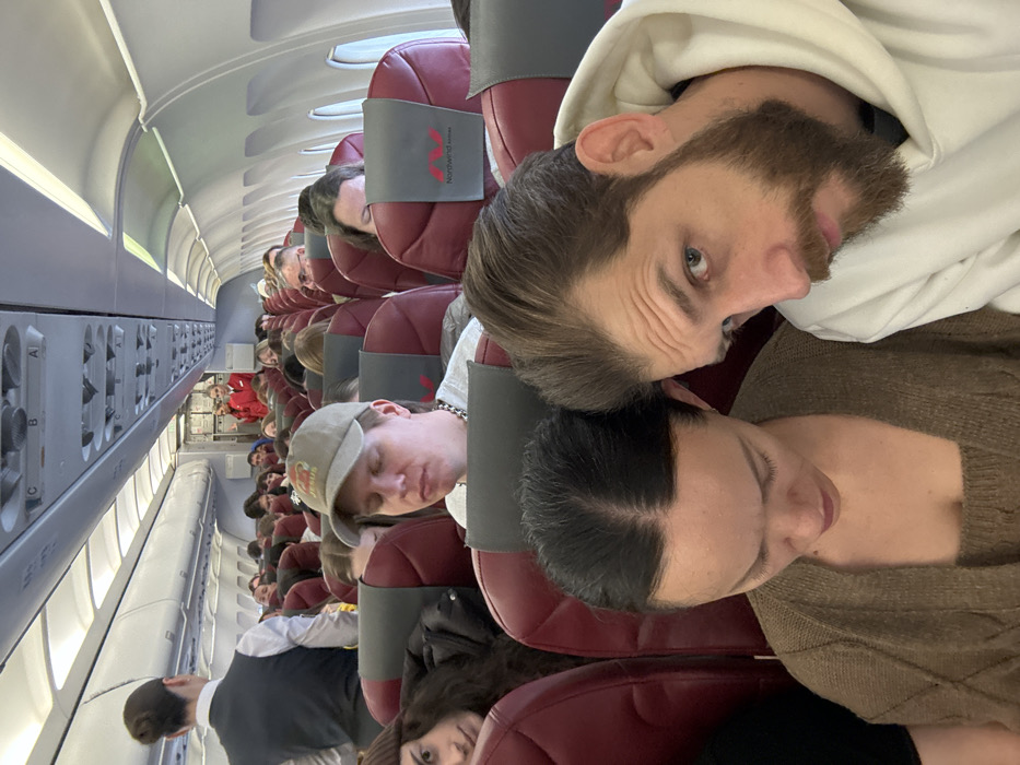 The scene depicts the interior of a commercial aircraft cabin, identified as a Nordwind Airlines flight by visible seat branding. The specified location for this event is Novogorelovo, Russia. The image captures a dense arrangement of passengers seated in dark red/maroon economy class seats with grey headrest covers, extending towards the front of the aircraft.
In the immediate foreground, a male with a brown beard and light eyes, wearing a white hoodie, is positioned on the right, looking directly towards the camera. To his left, a female with dark hair, wearing a brown knitted top, is looking downwards. Behind them, another female with long dark hair is partially visible, glancing towards the camera from the seat to the left. Central to the mid-ground, a male wearing a light-colored baseball cap with a "JESUS" graphic and a silver chain is seated with his eyes closed.
Further down the aisle on the left, a female flight attendant with her hair in a bun, wearing a dark vest over a white shirt, stands with her back mostly to the camera. In the distant background, near the front galley area, two additional female flight attendants in red uniforms are visible, one gesturing. Overhead bins and passenger service units are visible throughout the cabin. Windows on the right side of the aircraft reveal bright, ambient daylight. The cabin is filled with passengers, occupying most visible seats.