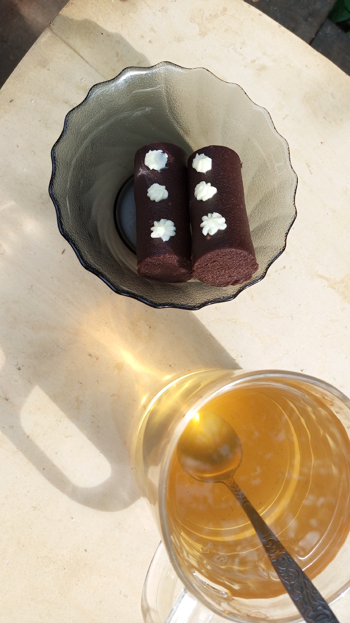 A top-down shot captures a light-colored wooden surface, partially shadowed by a glass of amber-colored liquid, presumably tea, with a metal spoon inside. A clear glass bowl with a rippled rim sits on the surface, containing two dark brown, cylindrical pastries, each decorated with four small, white, floral-shaped toppings. The location is Юдино, Russia. The scene suggests a simple outdoor tea break.
