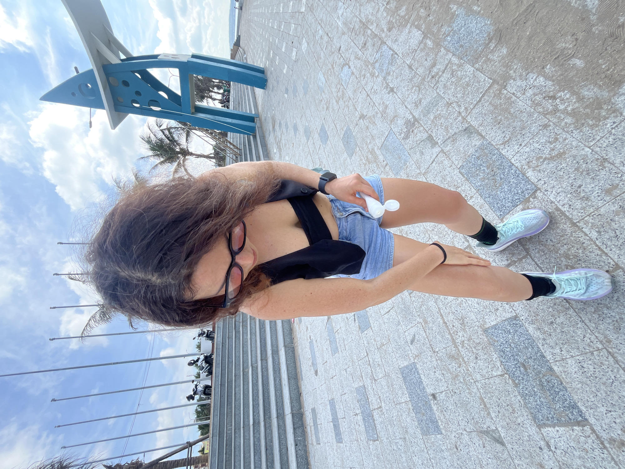 In Phan Thiết, Vietnam, a woman is standing on a tiled walkway outdoors. She is wearing a black crop top, denim shorts, black socks, and light blue and purple athletic shoes. The woman has long, dark, wavy hair and is wearing black-rimmed glasses. She is holding a small white tube in her right hand and appears to be applying cream or lotion to her leg with her left hand. A smartwatch is visible on her left wrist. In the background, there are tiered concrete steps leading up to a promenade with several flags on tall poles and some parked motorcycles. To the right, a distinctive blue and white abstract sculpture stands tall against a bright, cloudy sky with palm trees and other foliage visible. The walkway is made of large, light-colored tiles with darker gray rectangular accents.