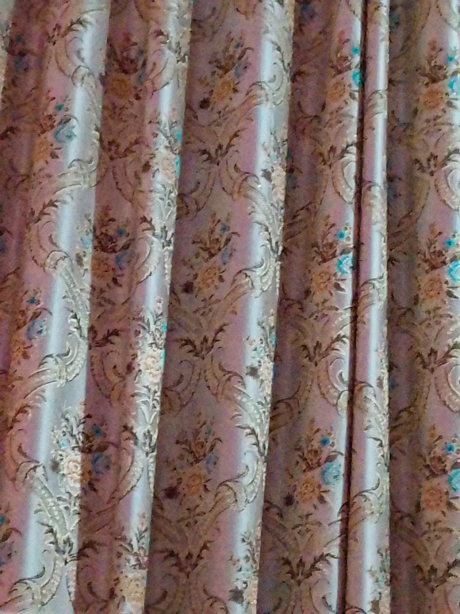 A close-up, full-frame shot showcases a textured curtain with a floral damask pattern. The fabric appears to be silk or a similar material, reflecting light and creating subtle highlights and shadows across its surface. The pattern features scrolling vines and stylized flowers in muted tones of cream, gold, and pink, with occasional pops of light blue. The curtain is draped, creating vertical folds that add depth and dimension to the image. The lighting is soft and diffused, suggesting an indoor setting. The image is devoid of any people or specific Mansehra landmarks, focusing solely on the textile's visual characteristics.