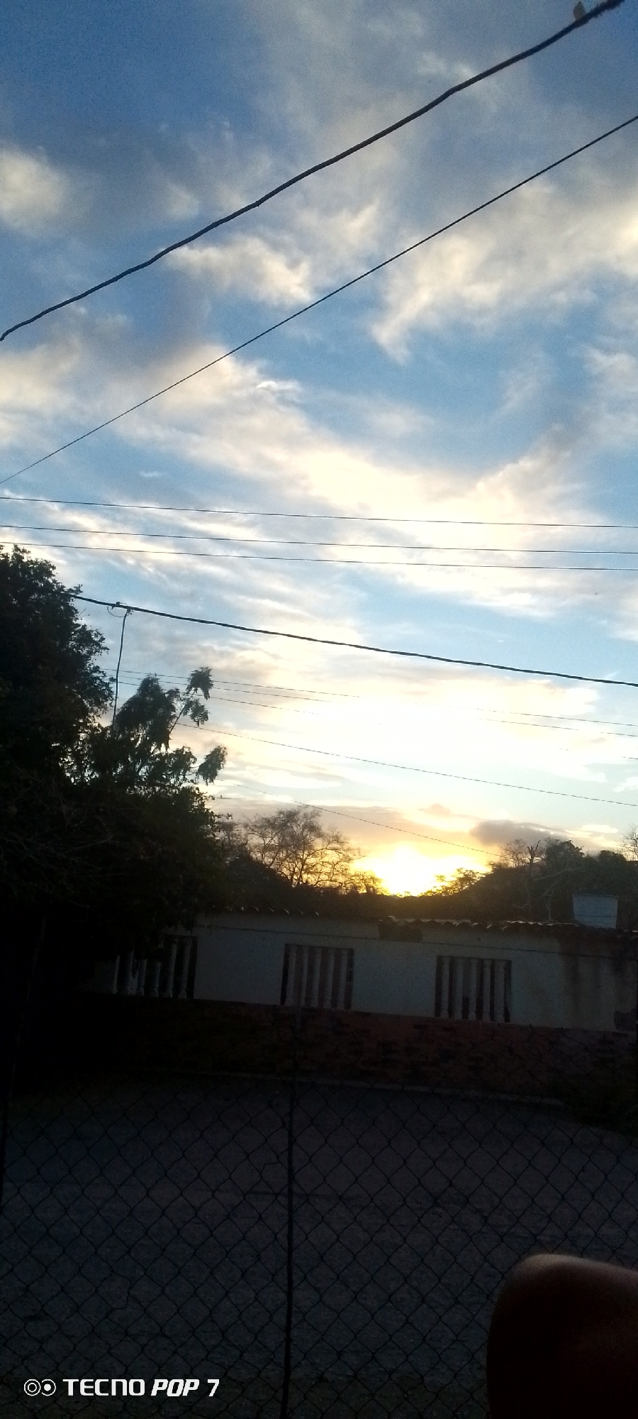 The photograph, taken with a TECNO POP 7 phone, captures a sunset scene in Altagracia, Venezuela. The sky is a gradient of blue and white clouds. High-tension power lines cross the top of the frame. A low building with barred windows is visible behind a chain-link fence. Trees are present on the left and behind the building, silhouetted against the setting sun. A finger is visible in the lower right corner.