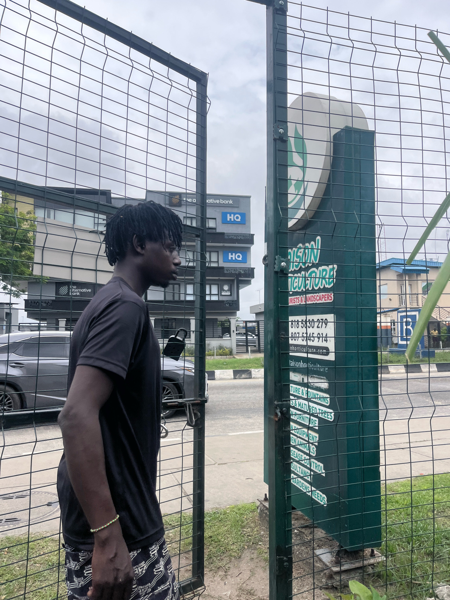 A young Black male with dreadlocks stands facing a green sign with white text behind a wire fence in Lekki Phase I, Nigeria. The man is wearing a black t-shirt and patterned shorts, with a beaded bracelet on his left wrist. The sign reads "TAISON HORTICULTURE" with smaller text below listing services like "FLOWER & FOUNTAINS", "GARDEN FURNITURE", and "PEST CONTROL". To the left of the sign is a building with "HQ" written on it. The background shows a gray sky and a street with cars. The overall environment appears to be urban with some greenery.
